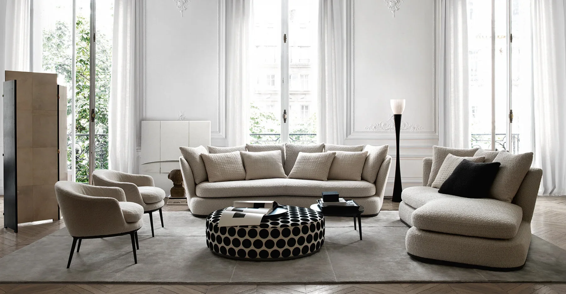 Living room with beige sofa, armchairs, black and white polka dot ottoman, and large windows with white curtains, bright natural light, and decorative wall molding.
