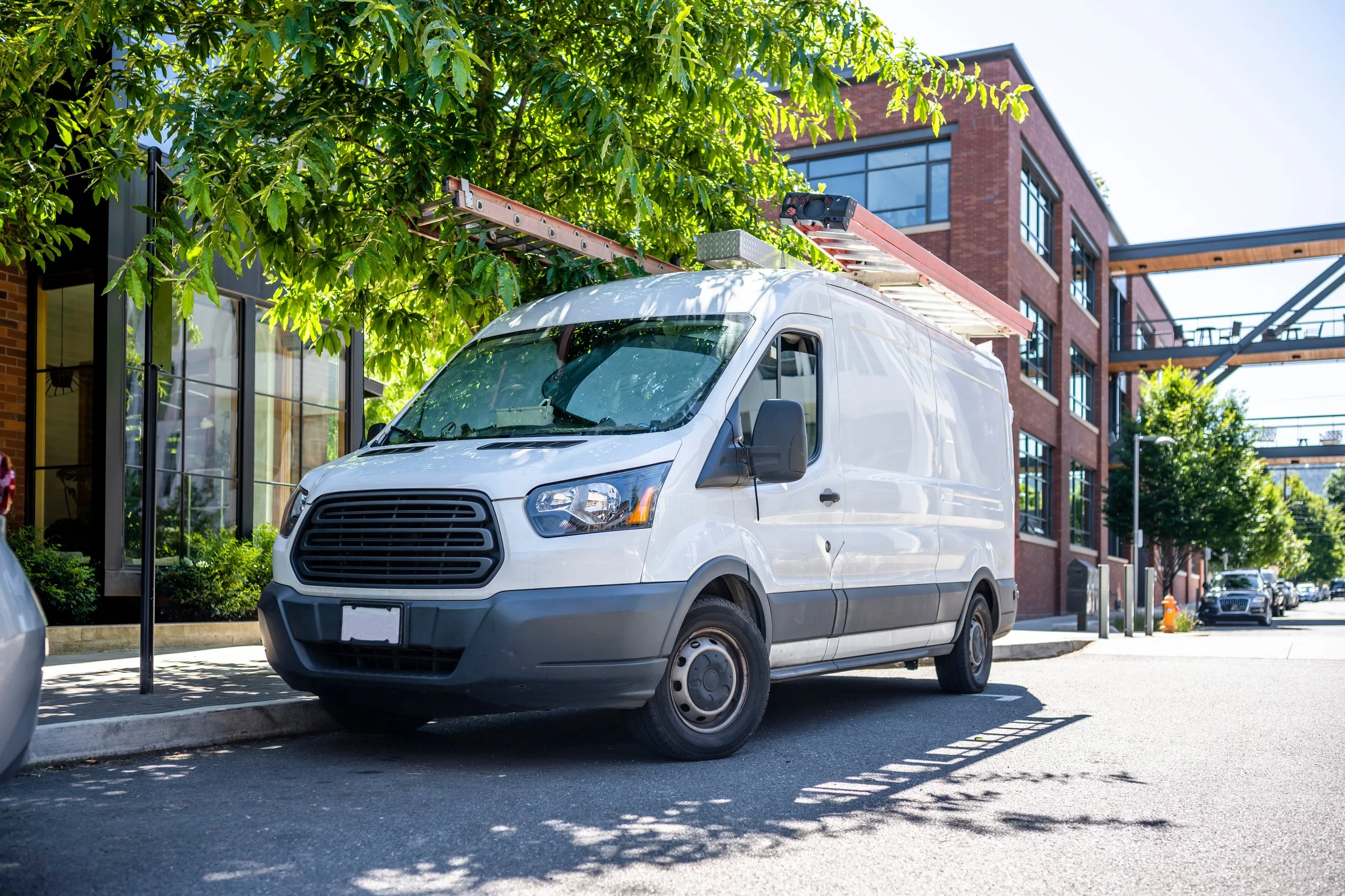 White utility van parked on the side of a city street under trees, with modern buildings, glass windows, and a fire escape in the background.