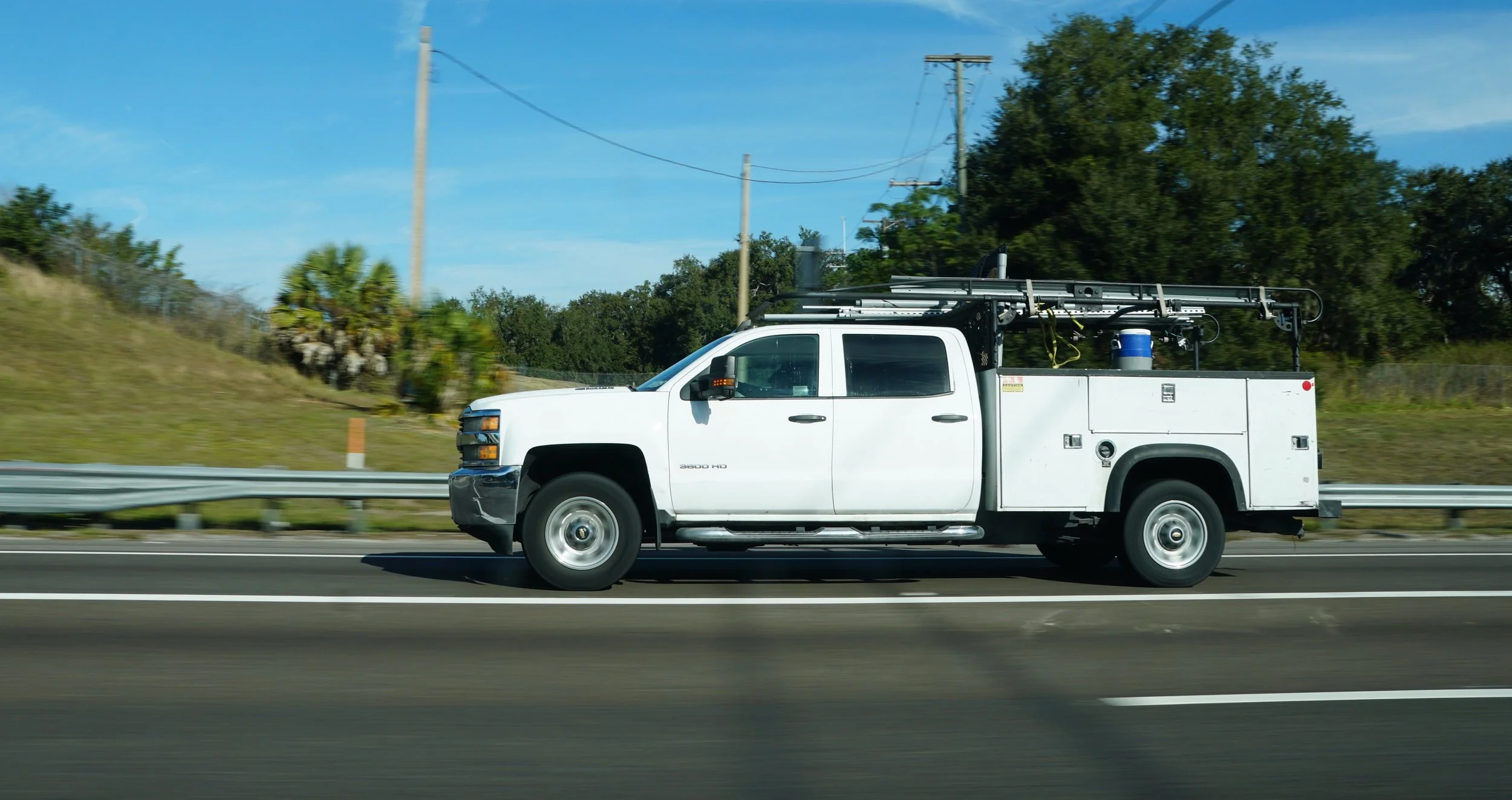 A white utility truck driving on a highway with trees and power lines in the background during the daytime.