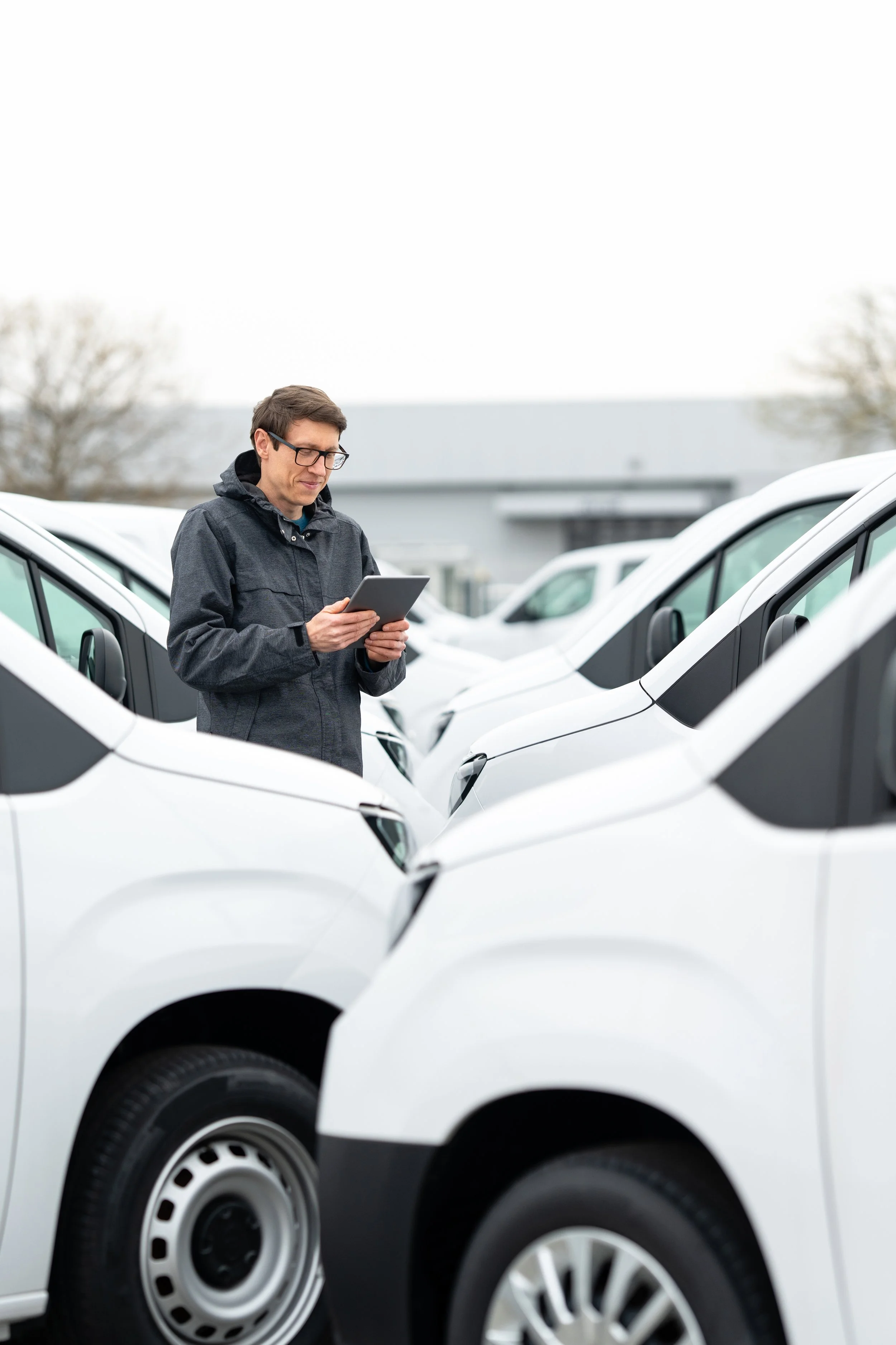 A man with glasses and a dark jacket is standing among white vans, looking at a tablet in an outdoor parking lot on a cloudy day.