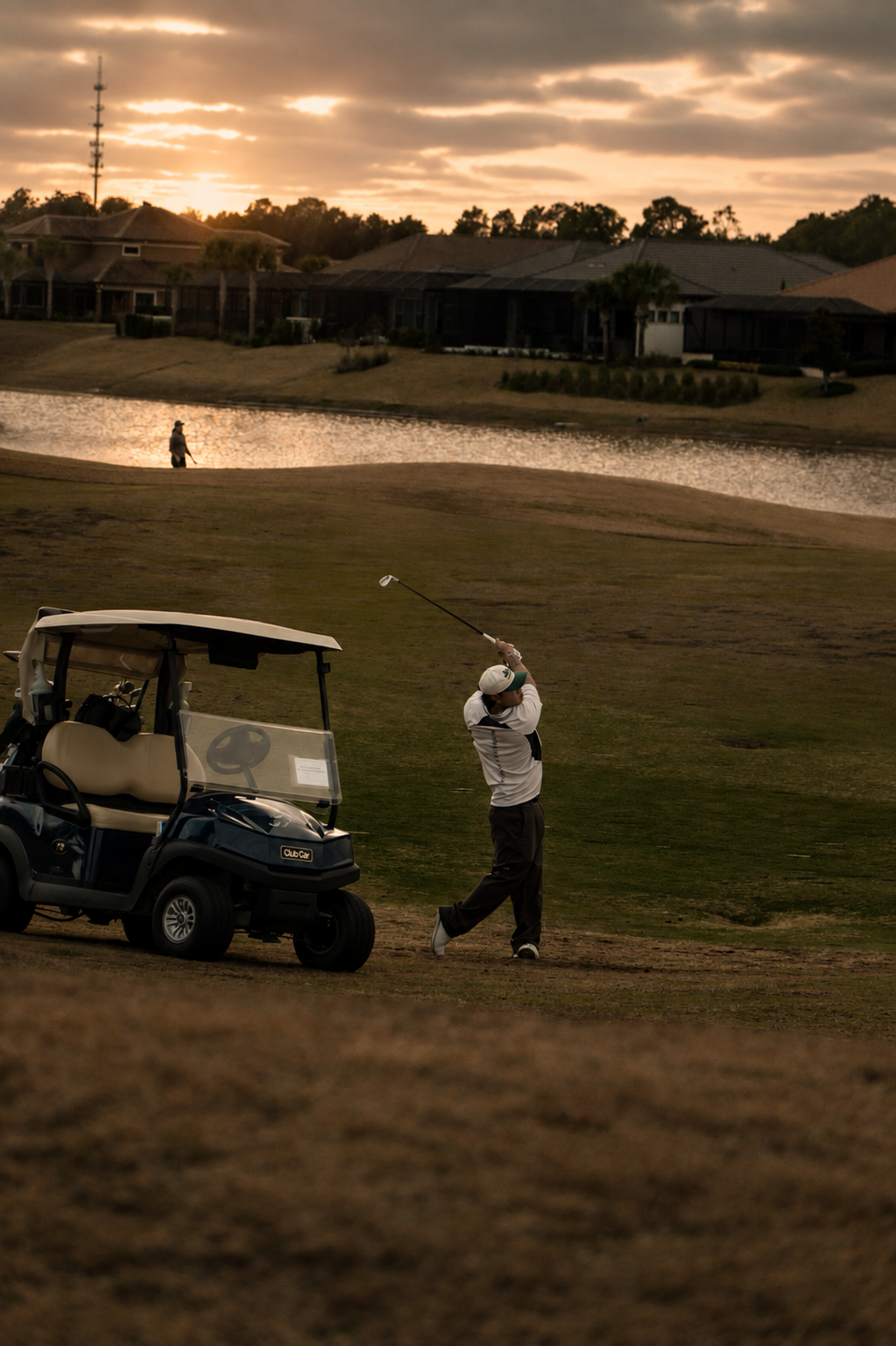 A person playing golf swinging a club near a golf cart at sunset with a lake and houses in the background.