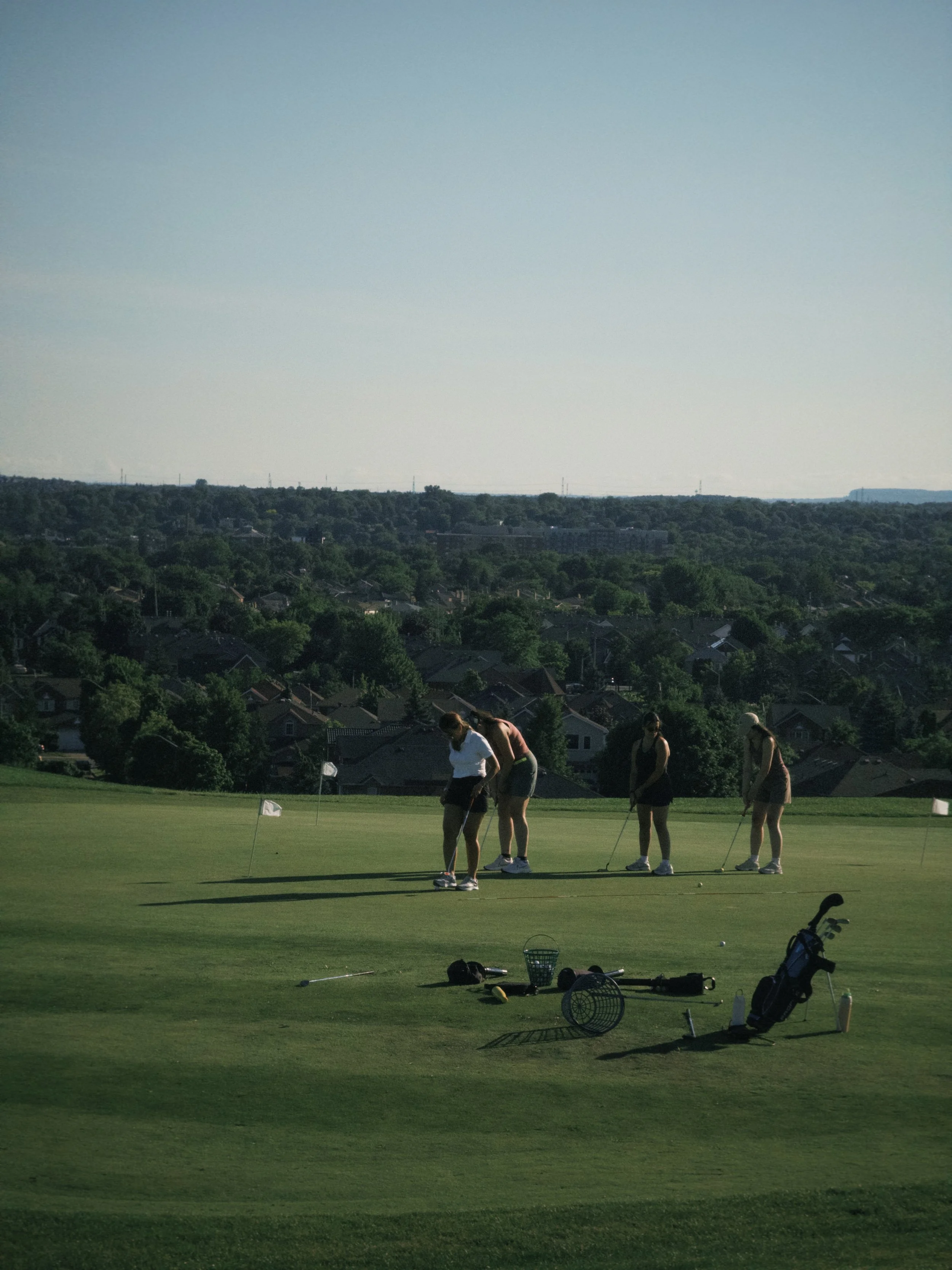 Four women playing golf on a green, with golf carts and equipment nearby, and a hilly landscape with trees and buildings in the background.