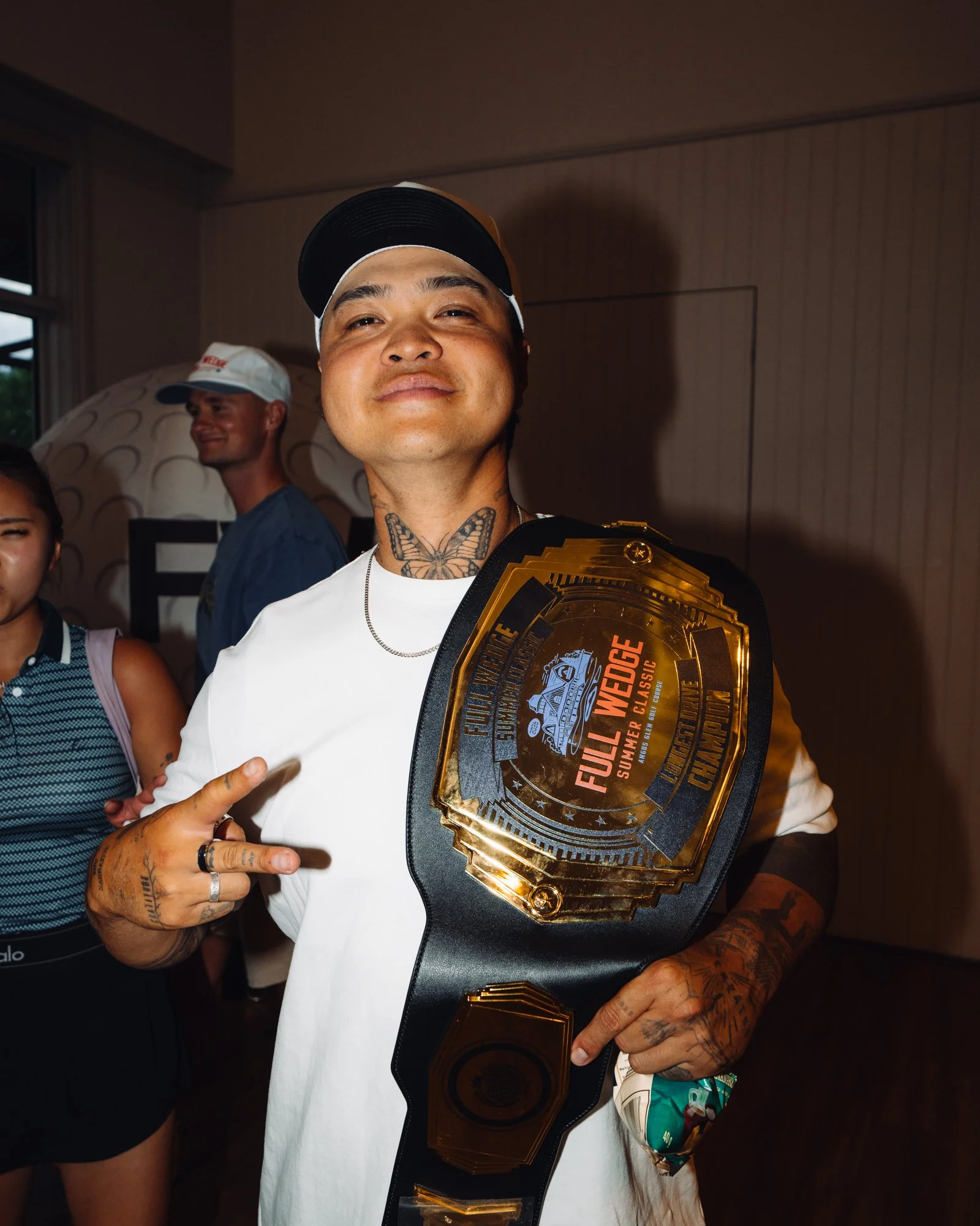 A man with tattoos on his neck and hands, wearing a black and gold championship belt, making a peace sign, wearing a white cap and shirt. Several other people are in the background.