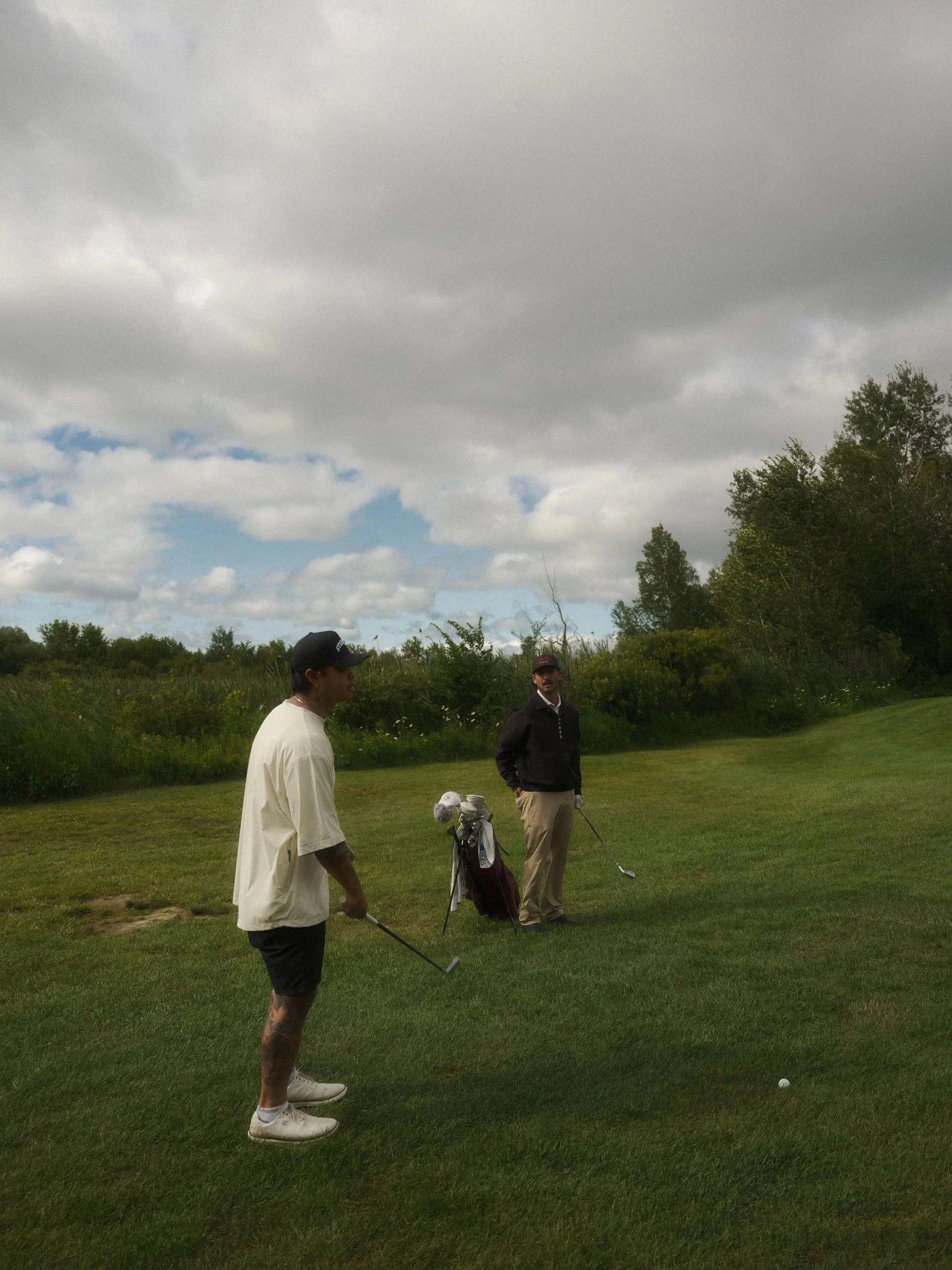 Two men are standing on a golf course, preparing to hit a golf ball, with one holding a golf club and the other standing nearby. There is a golf bag with clubs on the ground. The background includes green trees, grass, and a cloudy sky.