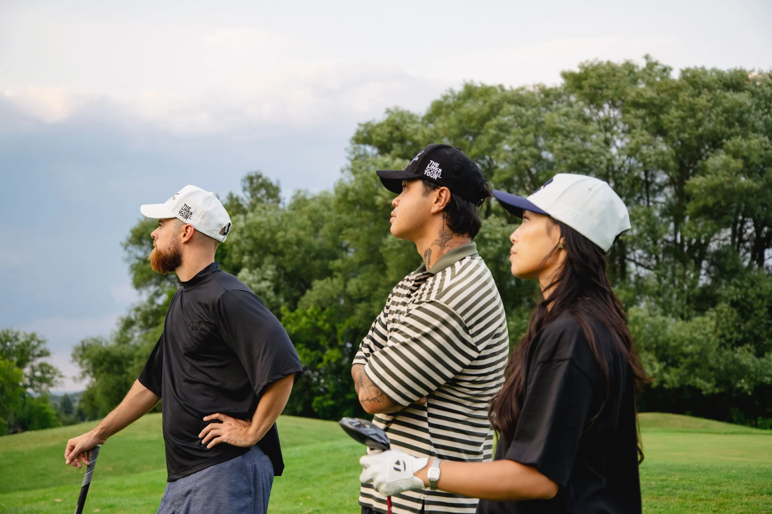 Three people standing outdoors on a golf course, wearing golf hats and casual clothes, with trees and a cloudy sky in the background.