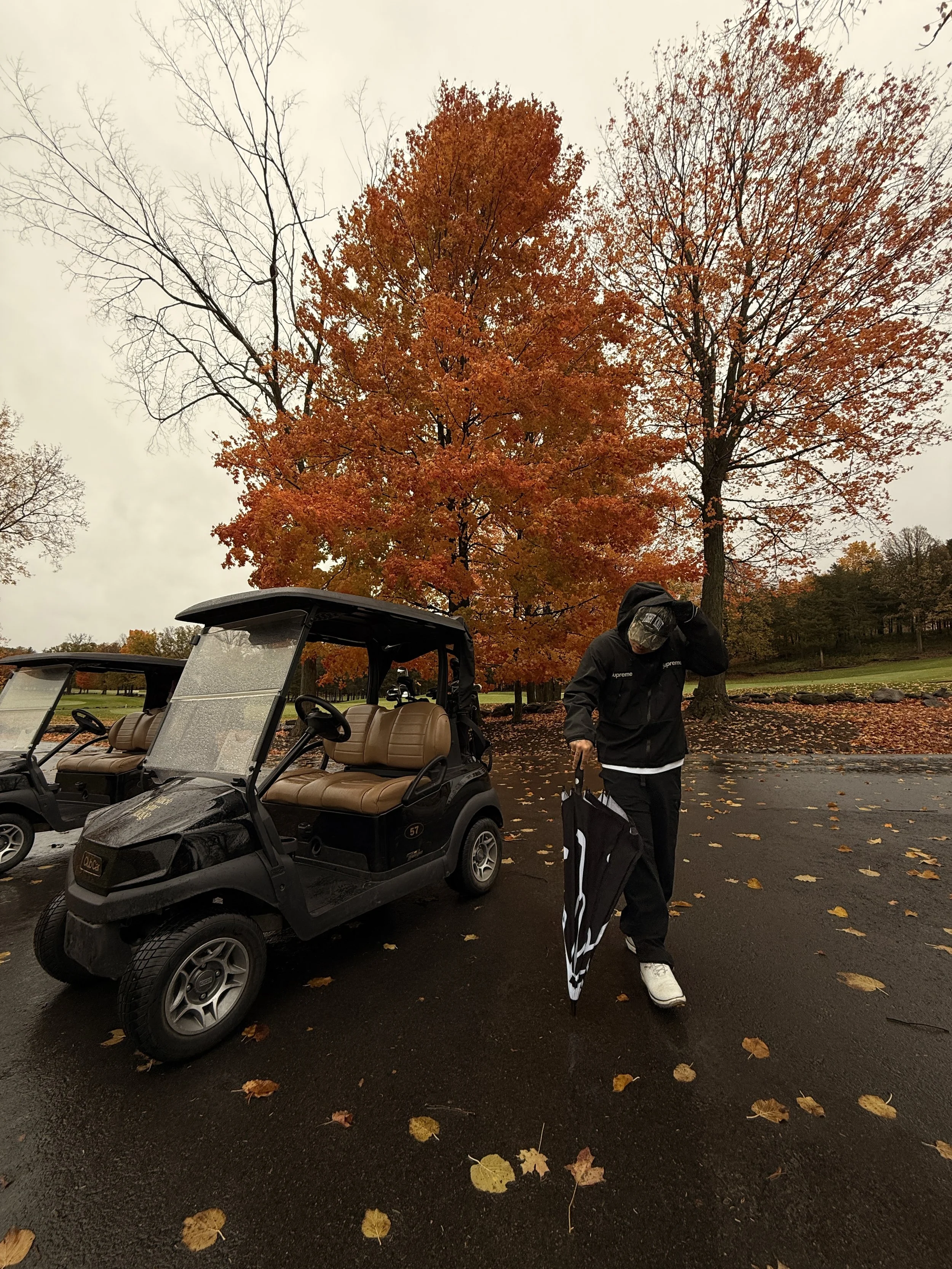 A person dressed in black walking with a golf bag near a black golf cart on a wet parking lot, with large trees with orange and red leaves and some fallen leaves on the ground, during cloudy autumn weather.