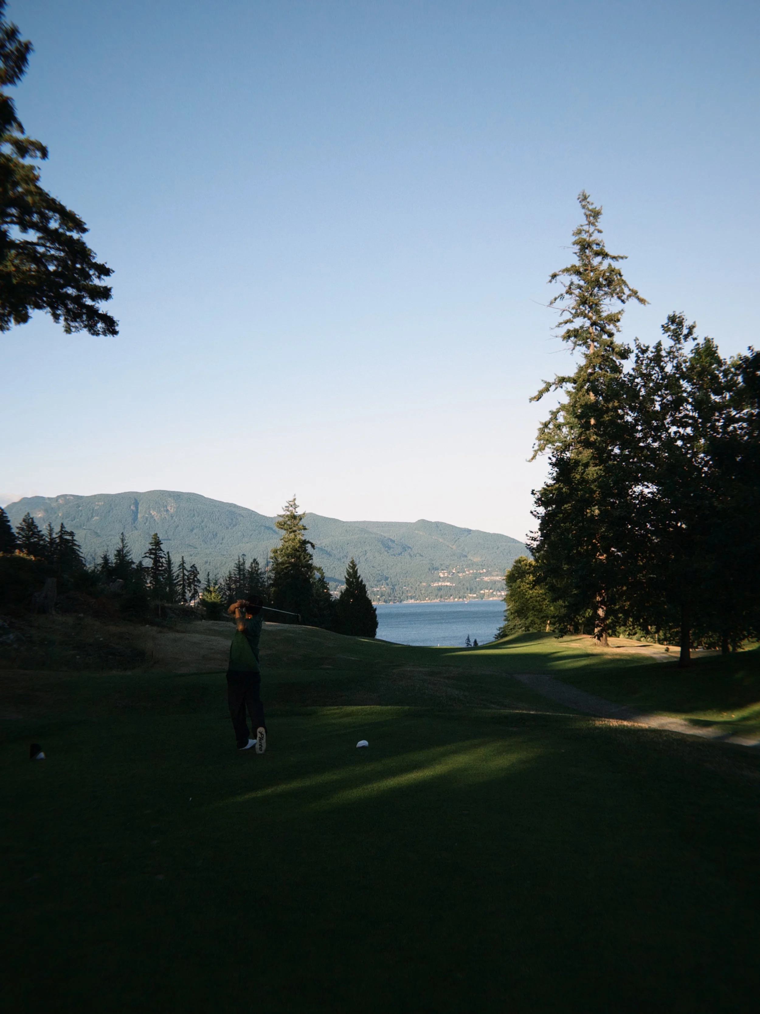 A person playing golf on a course with trees, mountains, and a lake in the background during sunset or late afternoon.