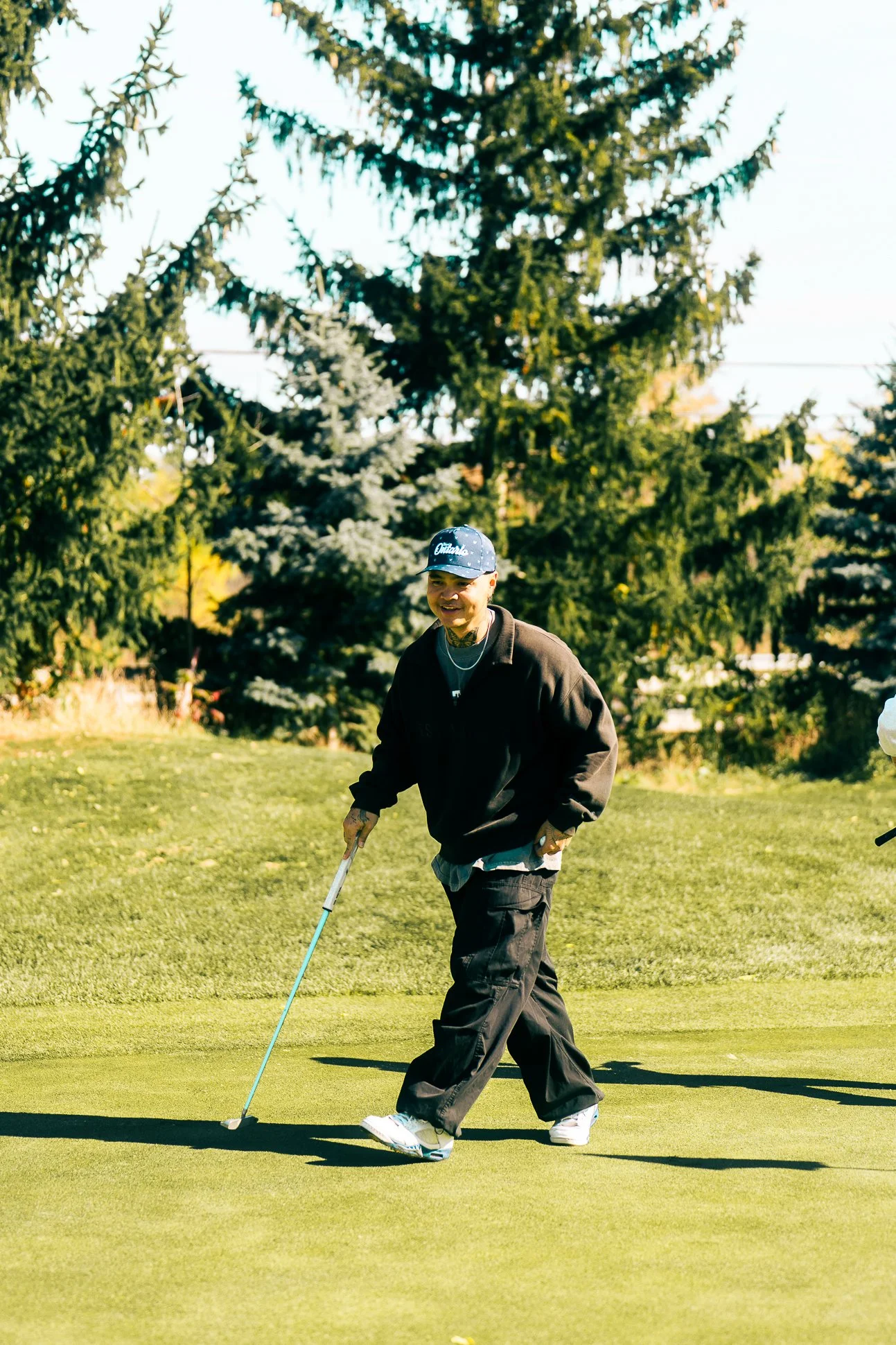 A person playing putt-putt golf on a grassy course, holding a golf club, wearing a black jacket, black pants, white sneakers, and a baseball cap with the word "Oakland" on it, with trees and a sunny sky in the background.