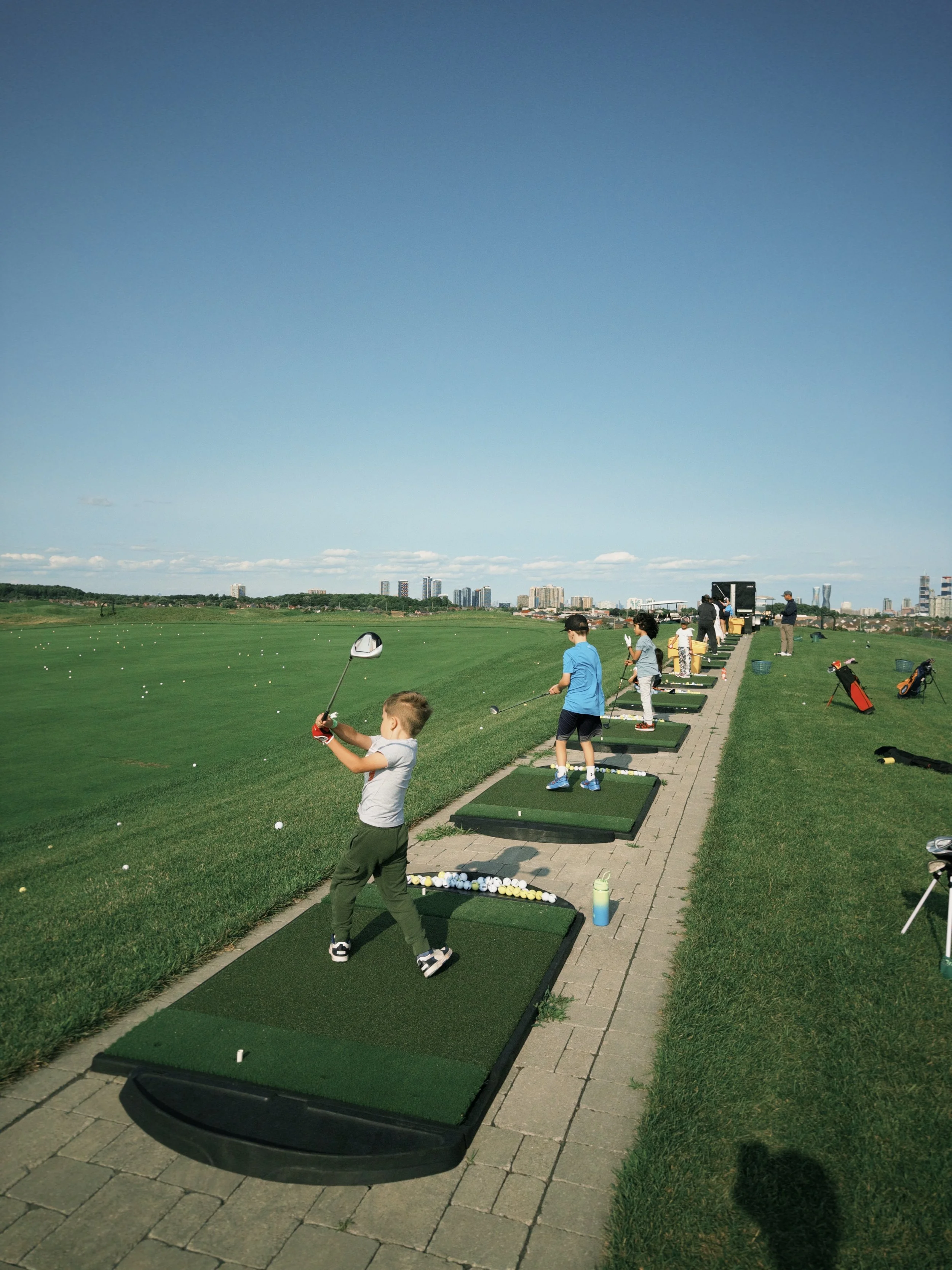Children practicing golf at a driving range with city skyline in the background on a clear, sunny day.
