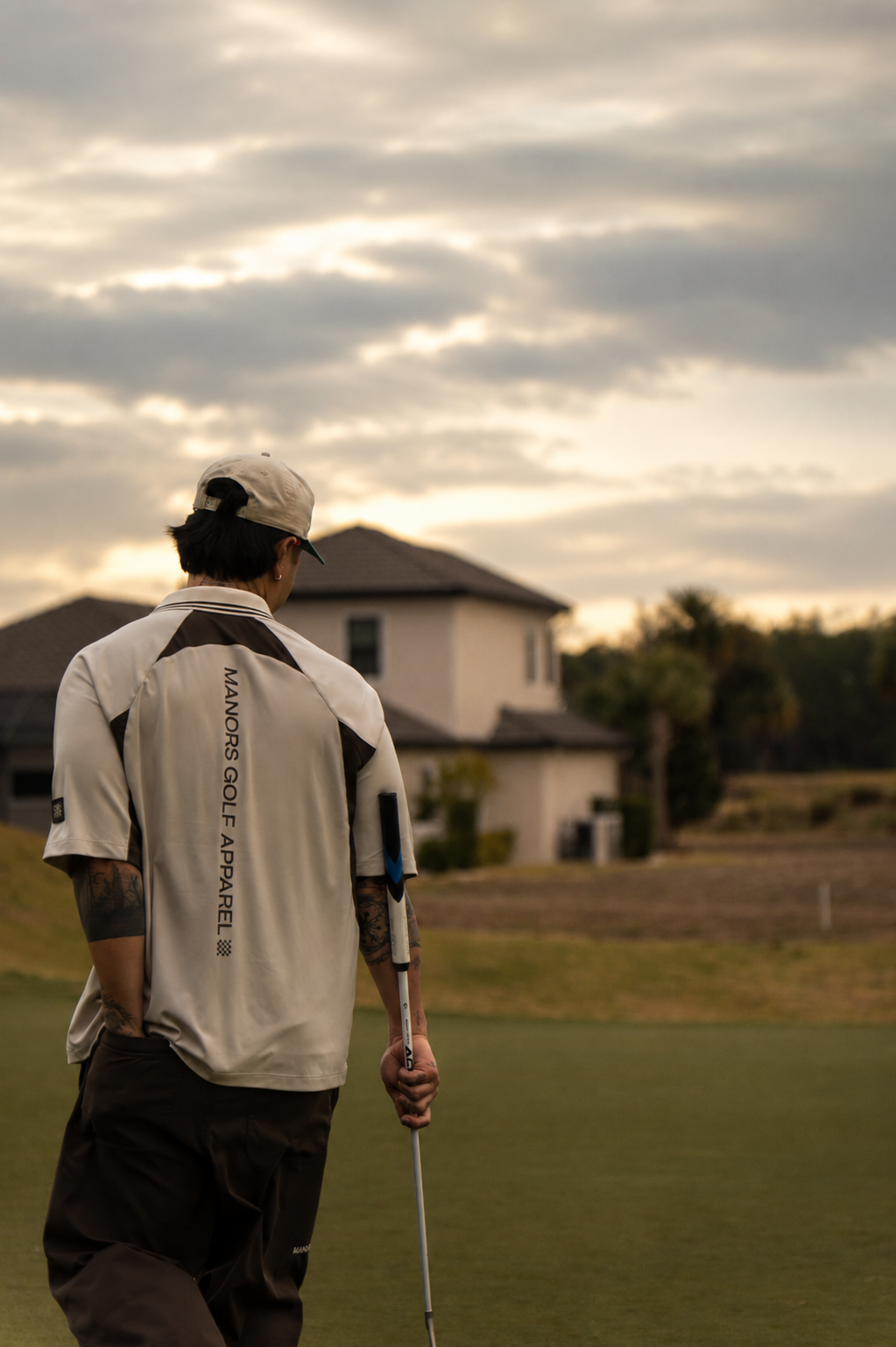 A person with tattoos on their arms, wearing a beige cap and a beige golf shirt with black accents, stands on a golf course holding a golf club, with a residential house and trees in the background during sunset or late afternoon.