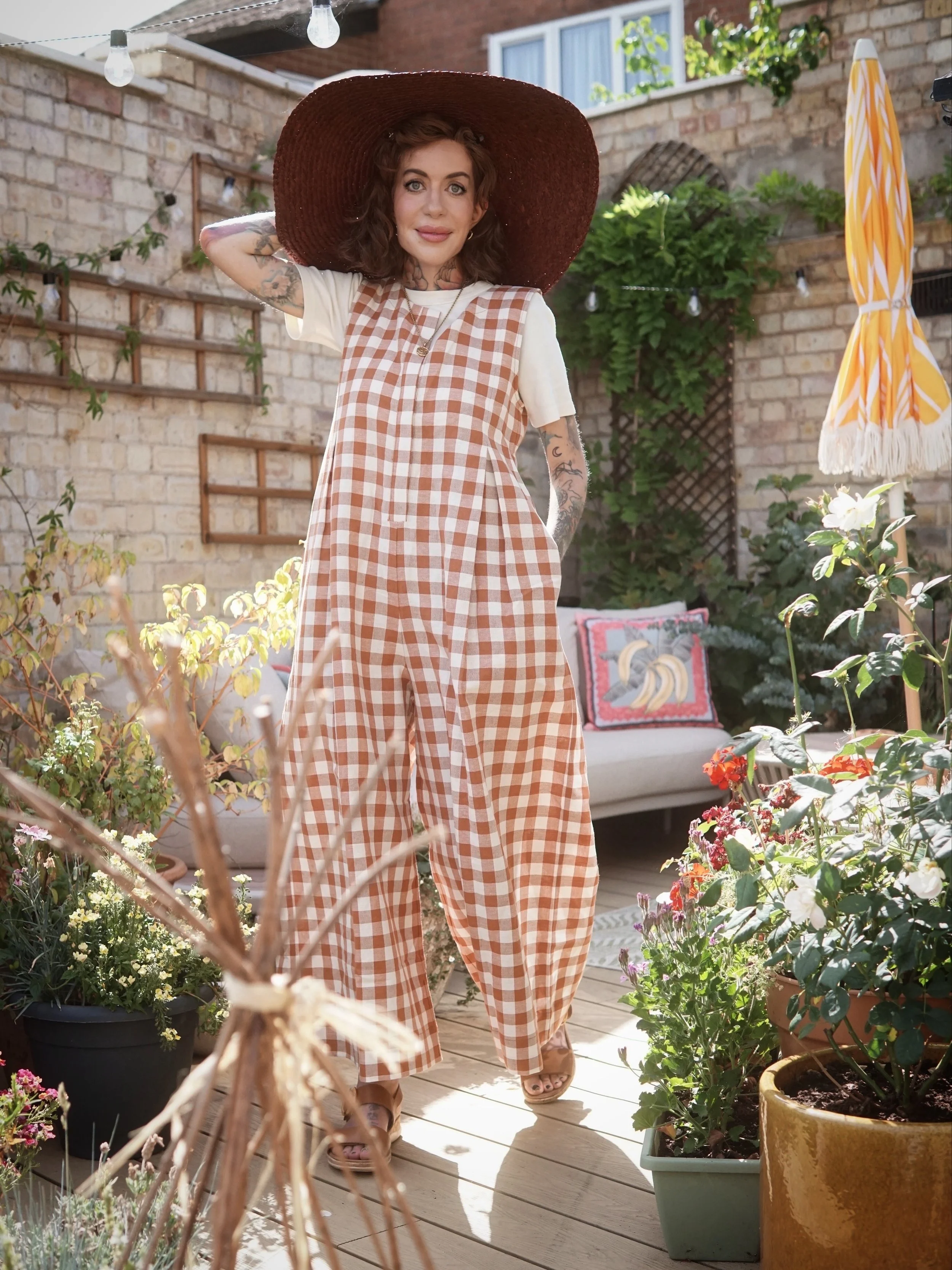 A woman standing on a wooden deck in a backyard garden with potted flowers, wearing a checkered jumpsuit and a large sun hat, smiling at the camera.