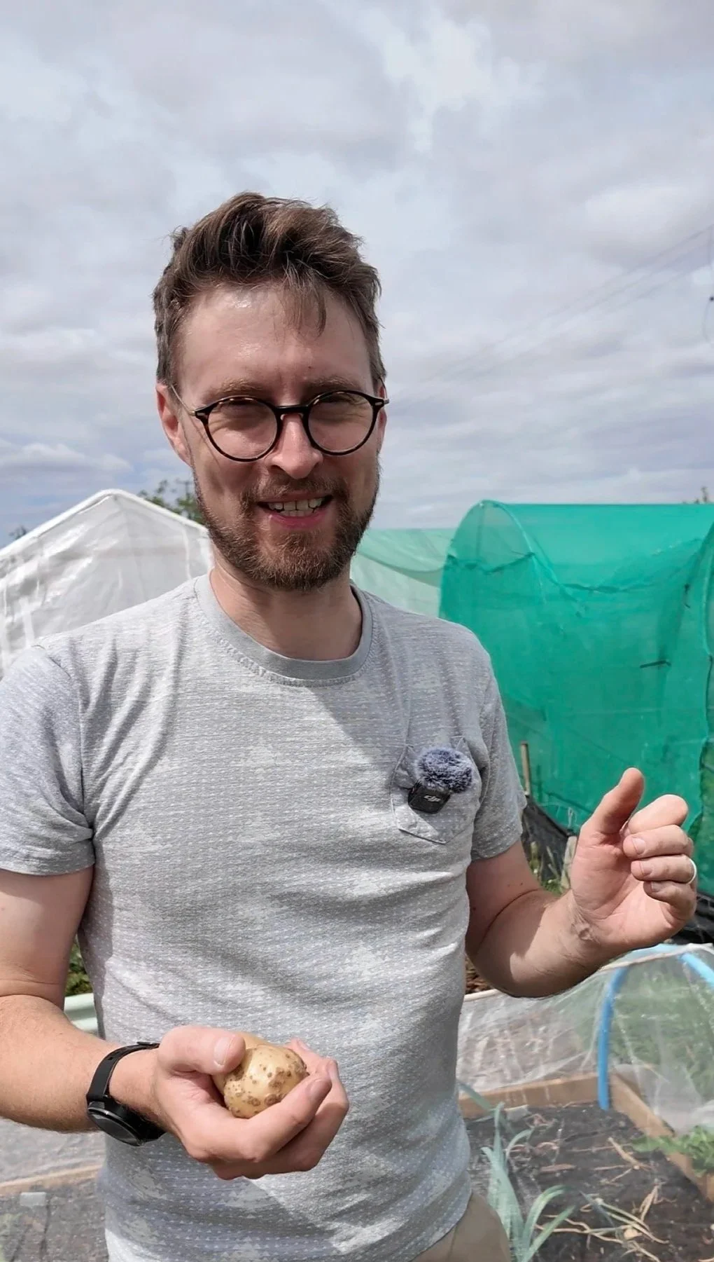 A man with glasses and a beard is holding a potato in his right hand, smiling and giving a thumbs-up, standing outdoors near greenhouses or plant covers on a cloudy day.
