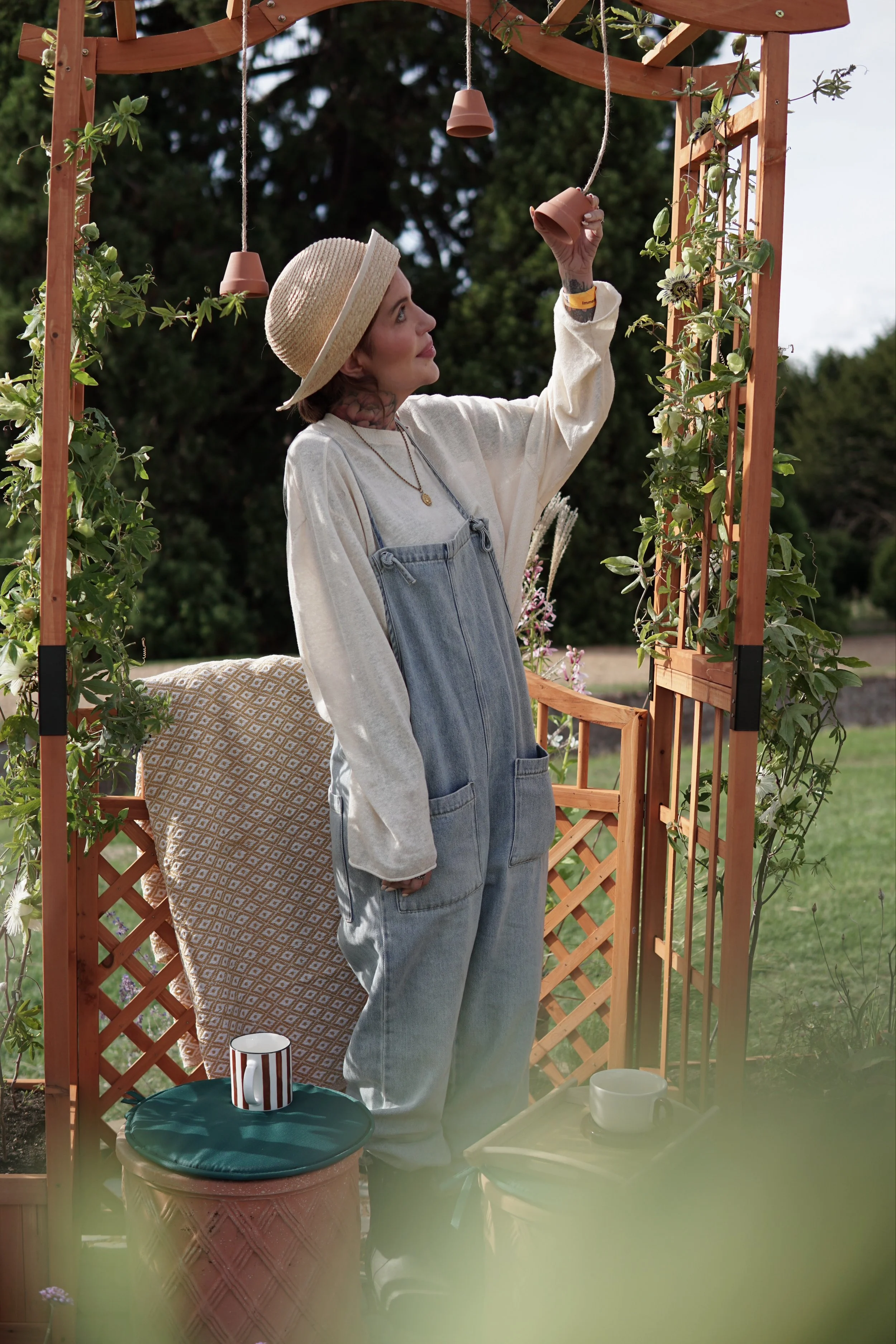 A woman in denim overalls and a wide-brimmed hat stands outdoors in front of a wooden garden arch decorated with green plants and flowers, holding a small brown ceramic cup. There is a table with a cup, a bowl, and other items, and a striped mug on a ceramic plant pot.