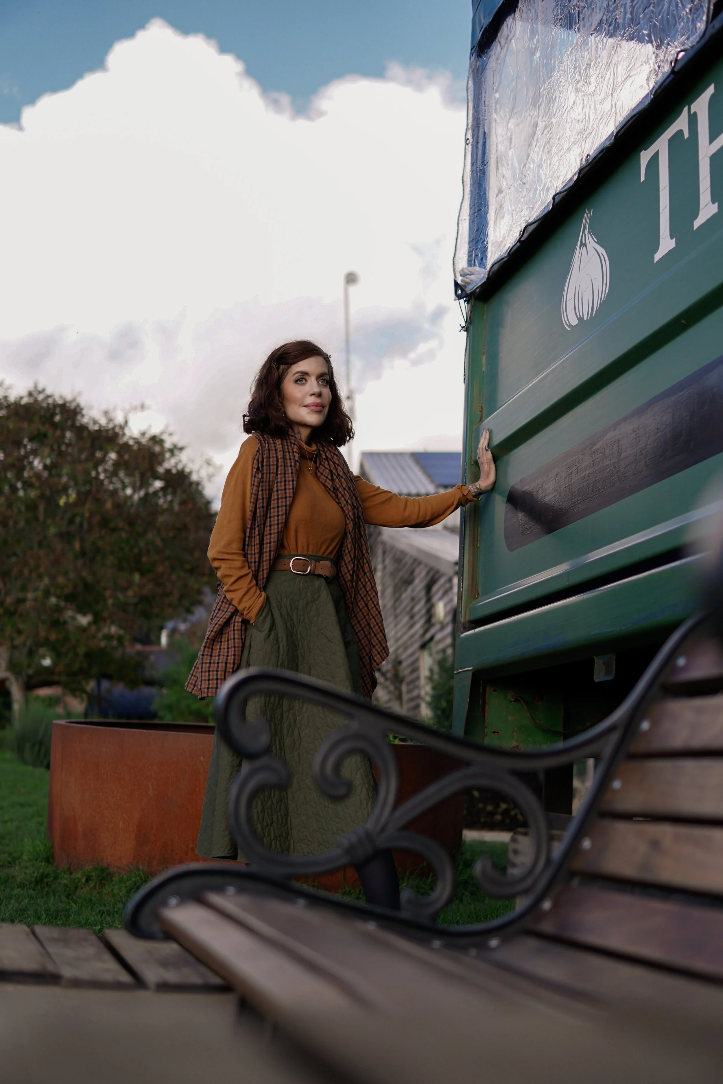 A woman with dark wavy hair and wearing a brown shirt and green skirt stands next to a blue food truck with a garlic illustration on it. She is touching the food truck with her right hand and looking away. There is a wooden park bench in the foreground and a large orange planter in the background. The sky is cloudy.