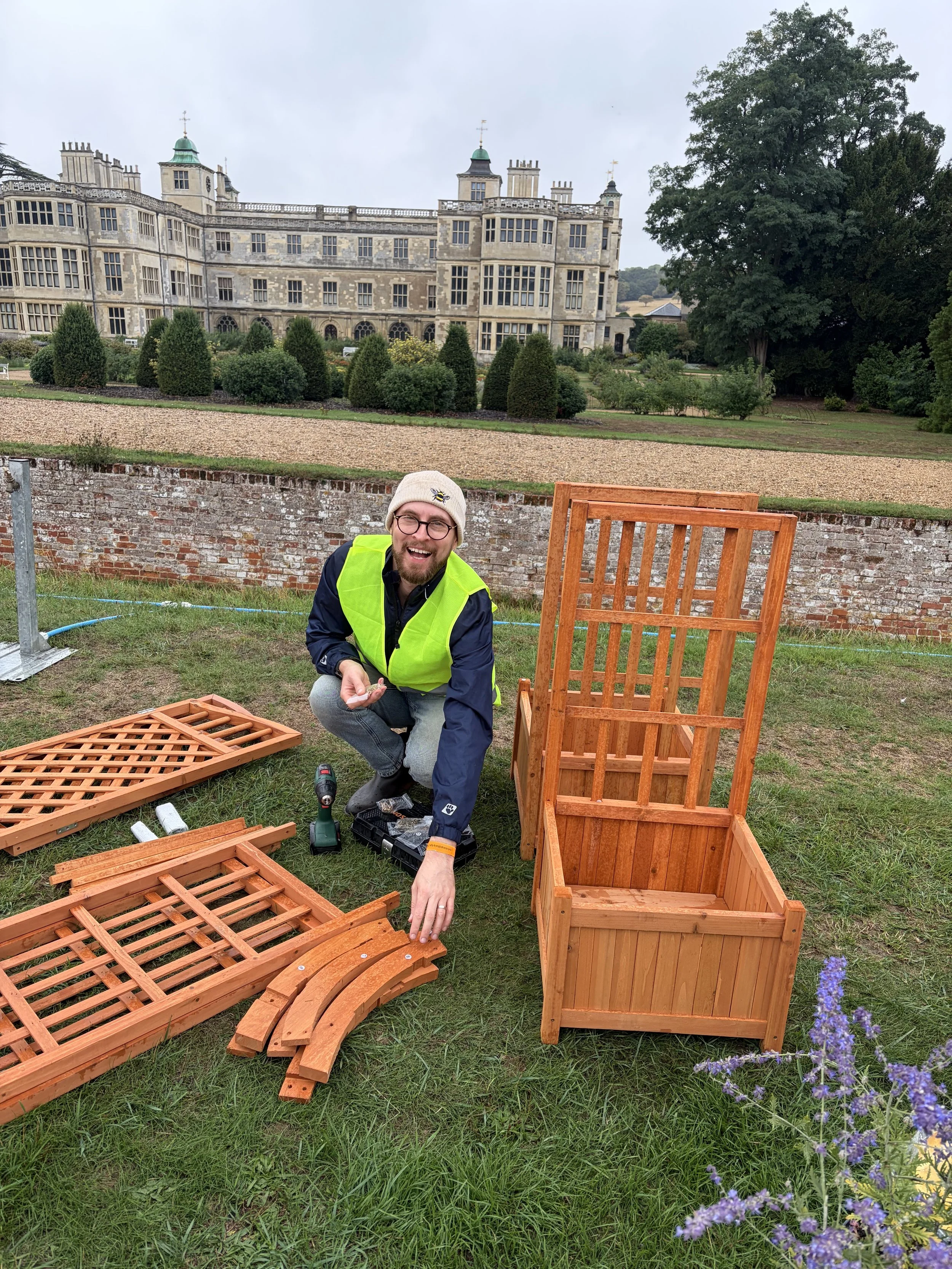 A man with glasses and a beard, wearing a knit hat and a yellow safety vest, crouching outdoors on grass, assembling wooden furniture with a drill, in front of a brick wall and a large historical building with numerous windows and towers in the background.