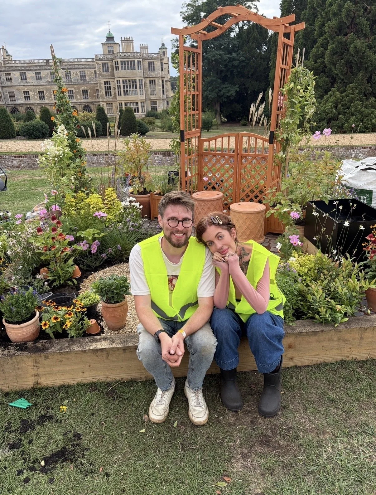 A smiling man and woman with bright yellow safety vests sitting on a garden bed surrounded by colorful flowers and plants, with an ornate large mansion in the background.