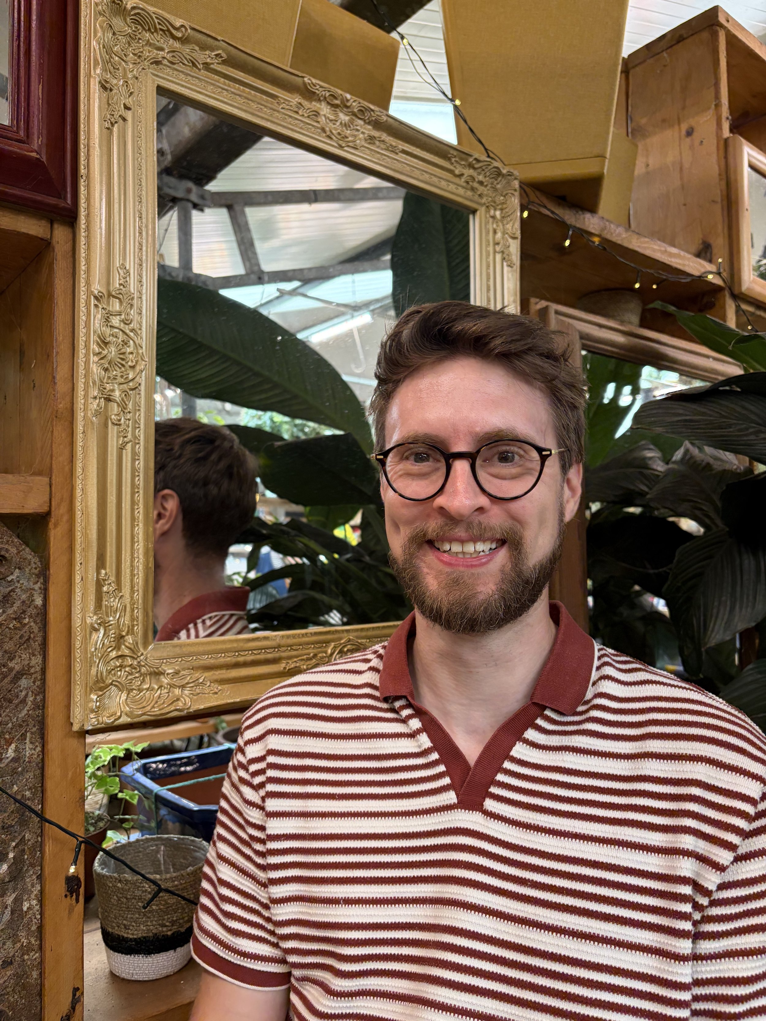 A smiling man with glasses and a striped shirt standing in front of a gold-framed mirror, with plants and wooden shelves in the background.