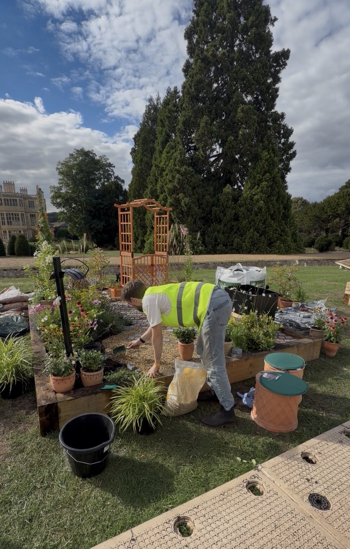 Person in a yellow safety vest gardening in a flower bed in a park or garden area, with trees and a building in the background under a partly cloudy sky.