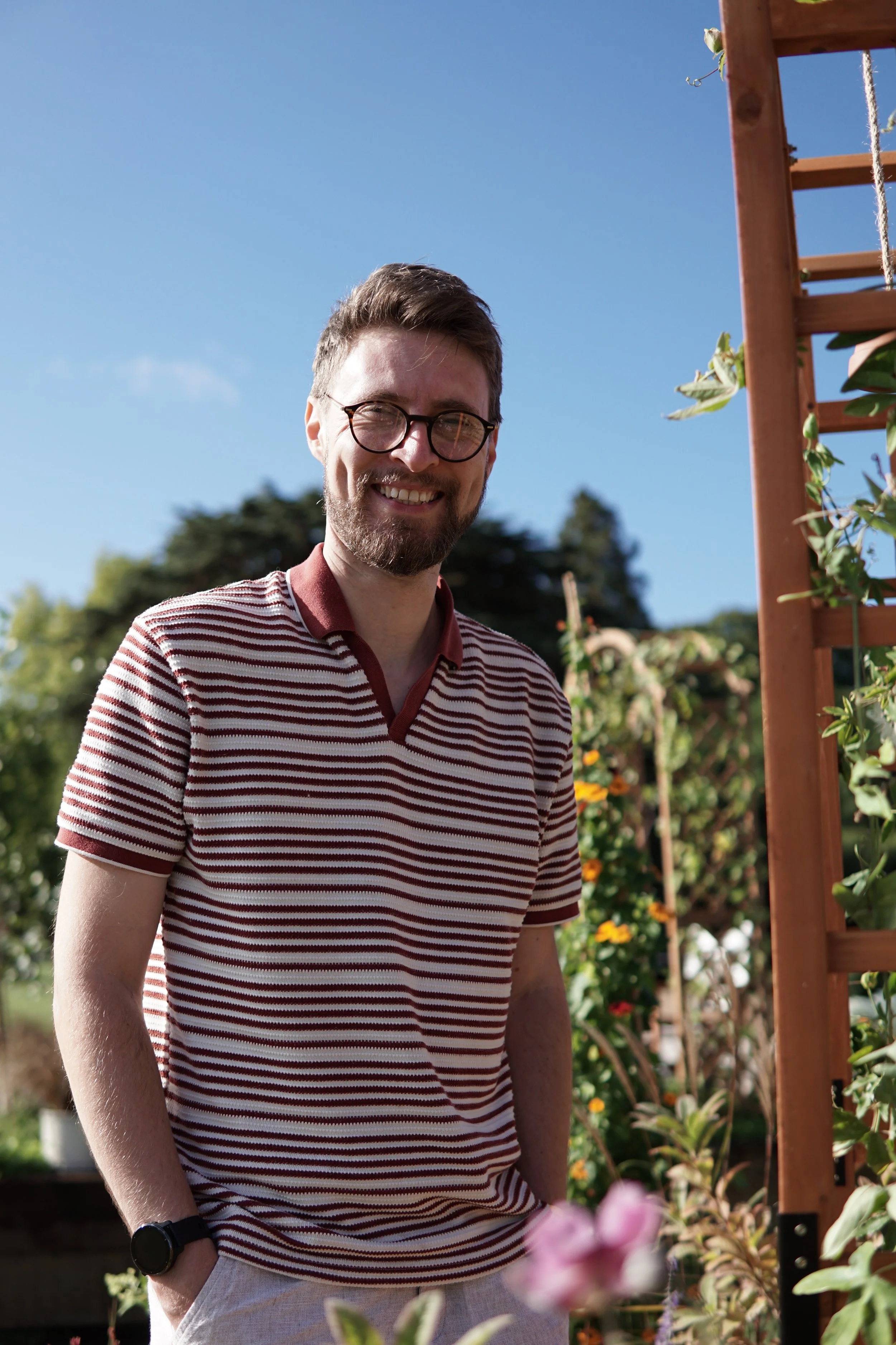 A smiling man with glasses and a beard, wearing a red and white striped polo shirt and a black watch, stands outdoors in a garden with colorful flowers and green foliage under a clear blue sky.