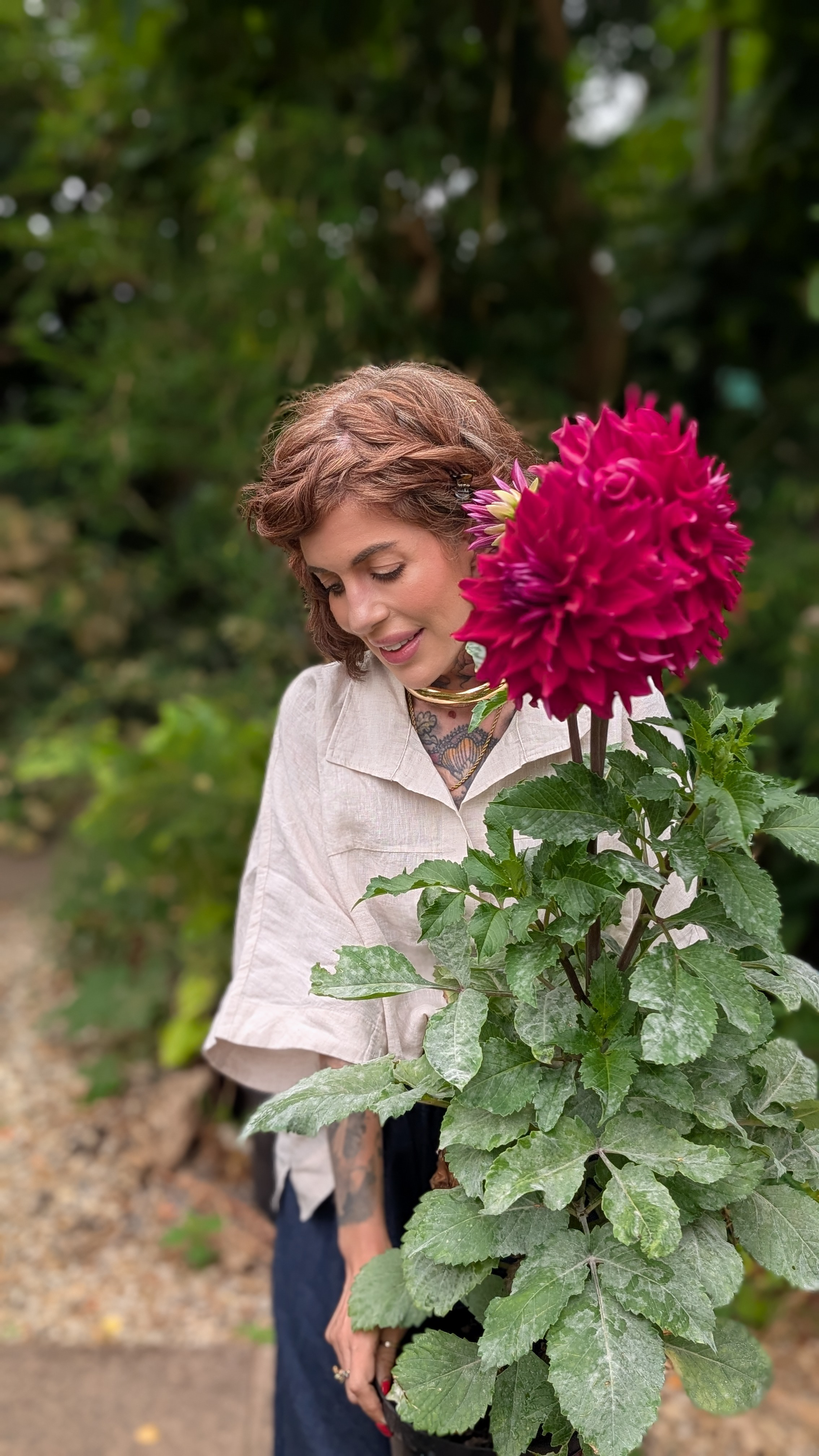 A woman with short brown hair, tattoos, and a beige blouse holding a potted plant with large pink flowers, standing outdoors with greenery in the background.