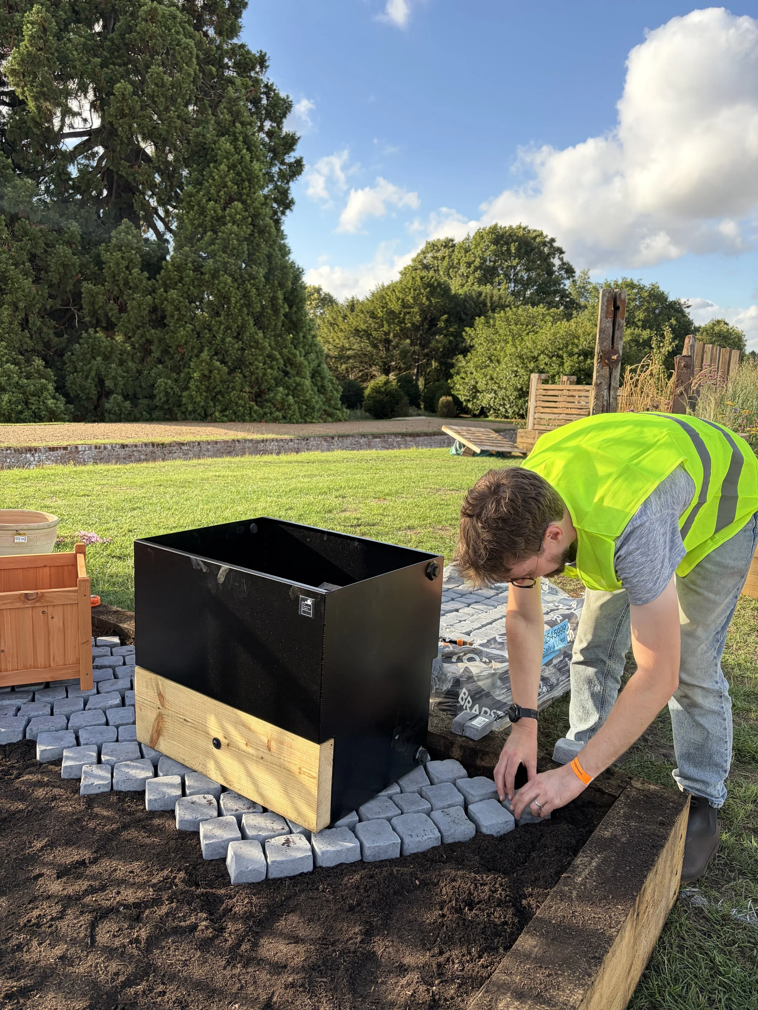Person installing paver stones in a garden bed outdoors on a sunny day, surrounded by green trees and a blue sky.