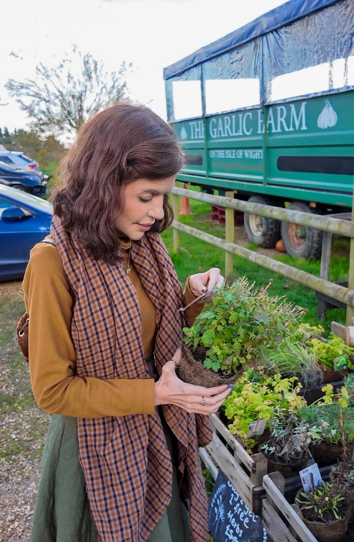 A woman with brown hair, wearing a brown checkered scarf and a tan coat, holding a small potted plant, standing outdoors near a display of various plants for sale at a farm or plant stand.