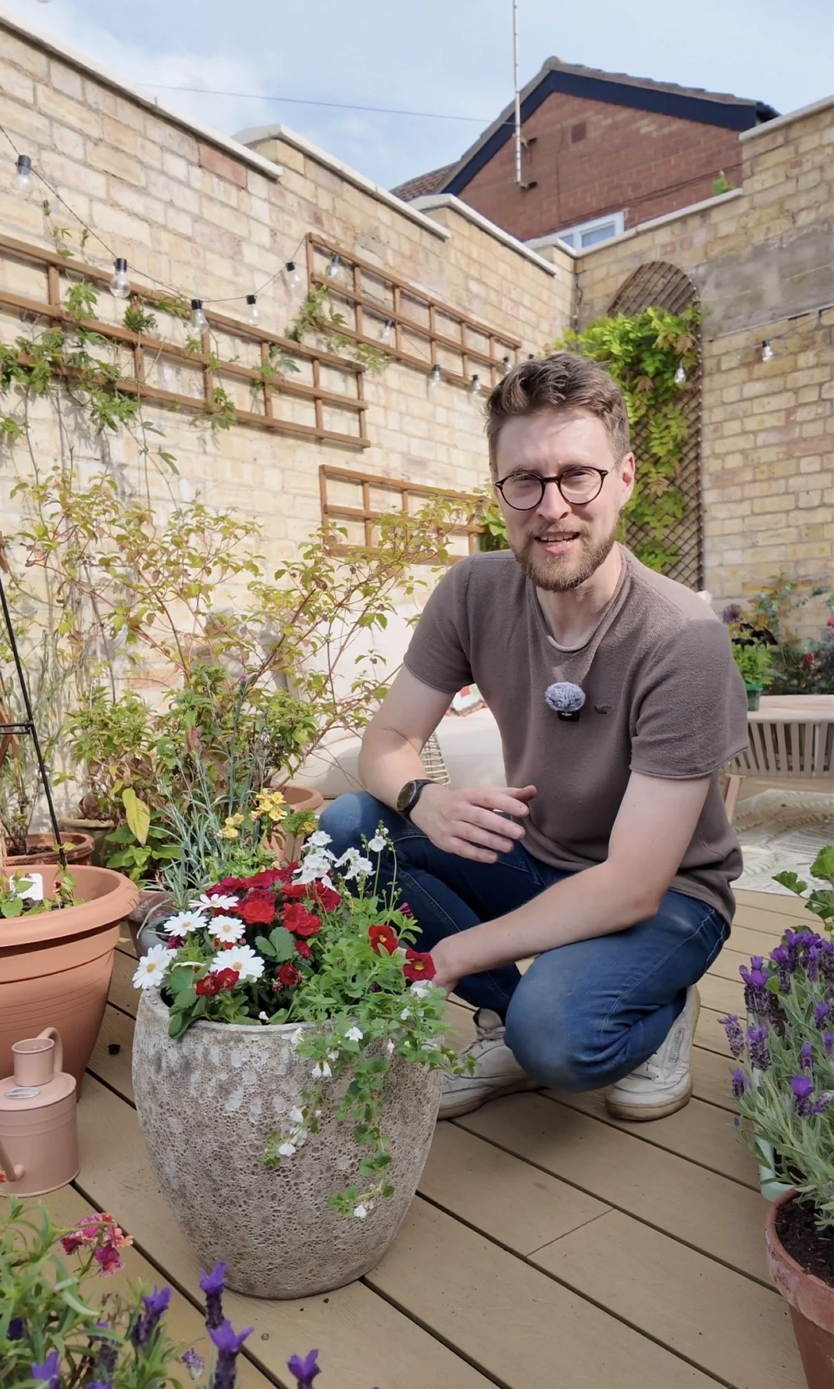 A man with glasses, a beard, and brown hair kneels on a wooden deck surrounded by potted flowers, including daisies and purple and red blooms, in a fenced backyard with a brick wall and house in the background during daylight.