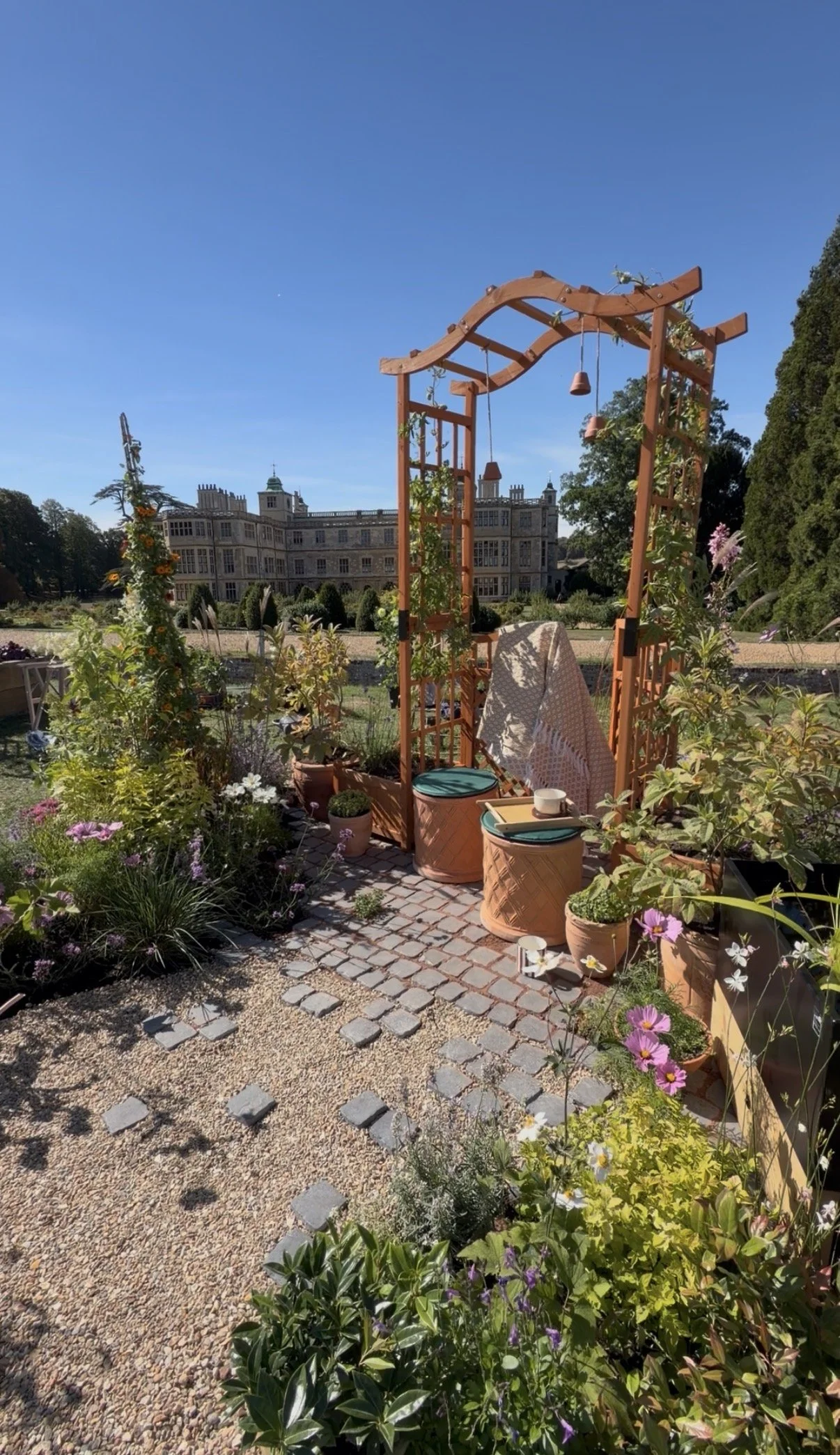 A garden scene with a wooden archway decorated with hanging bells and plants. There are potted plants, flowers, and a cobblestone pathway leading towards a large, historic building in the background. The sky is clear and blue.