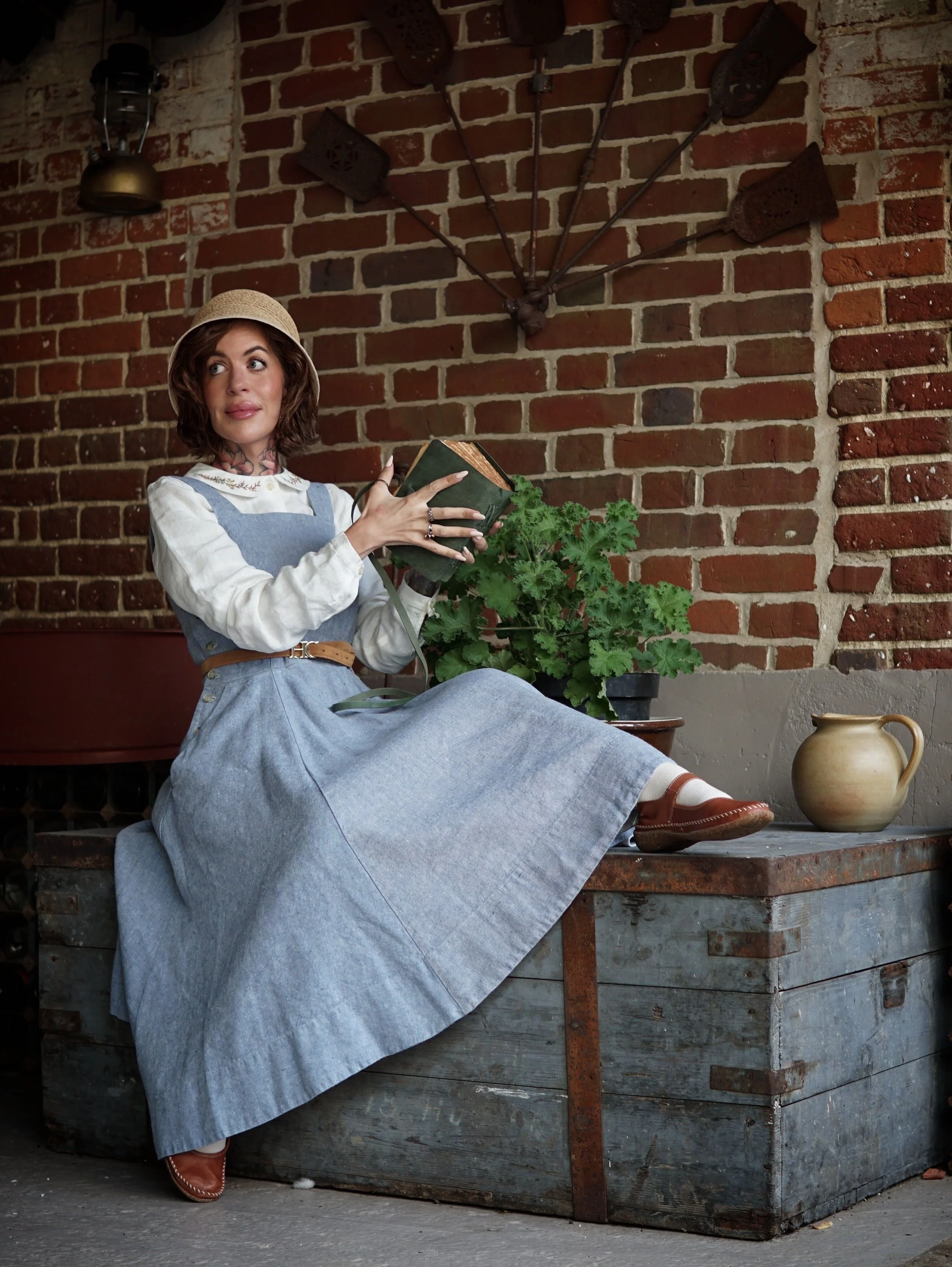 A woman with short, wavy brown hair wearing a straw hat and vintage-style clothing sits outdoors against a red brick wall. She holds a book in her right hand, relaxing with her left arm resting on her lap. Behind her is a large potted plant, and to her right is a rustic wooden table with a clay pitcher on it. A decorative metal umbrella and a lantern are mounted on the brick wall above her.