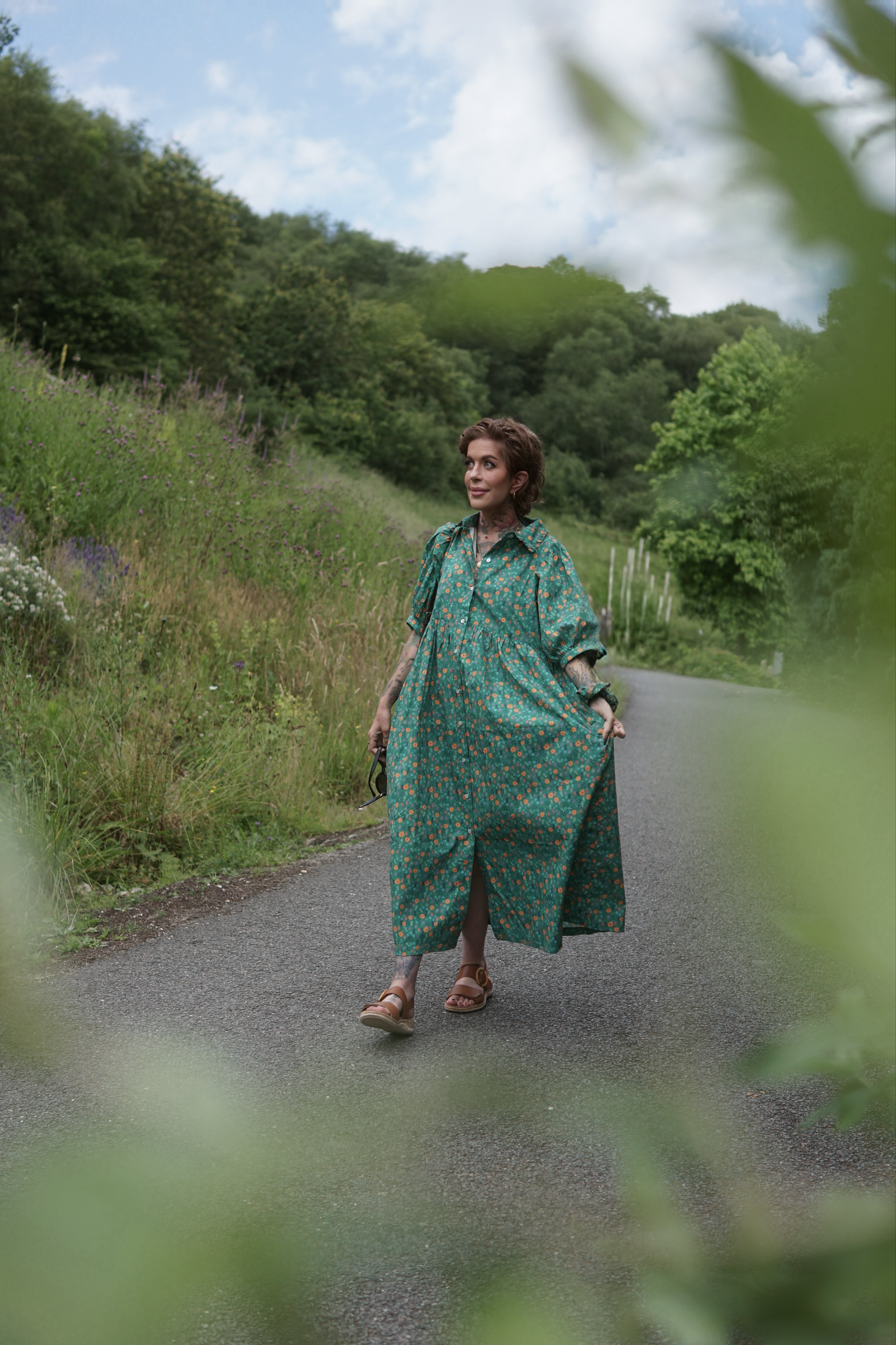 Woman in a flowing teal dress with orange floral pattern walking on a rural road surrounded by lush green trees and shrubs, with a cloudy sky overhead.