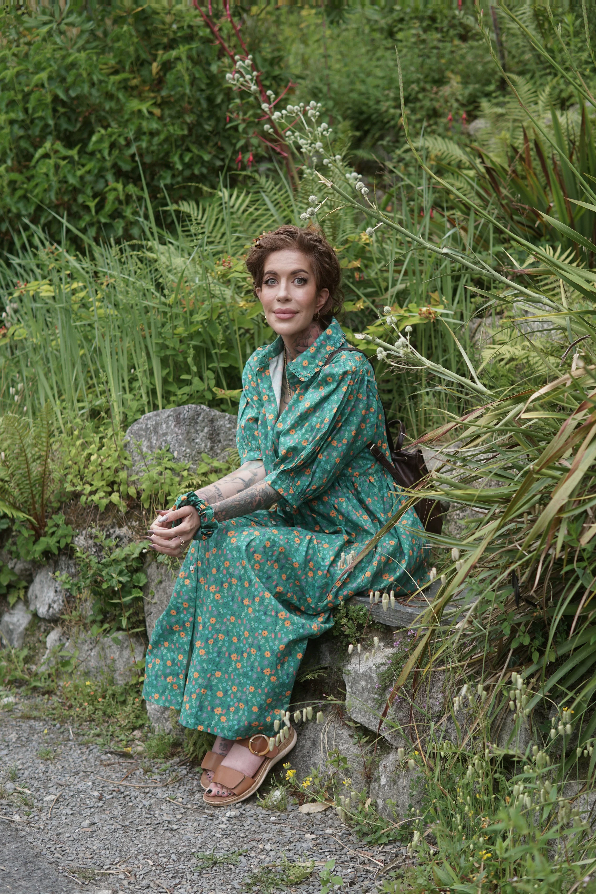 A woman with tattoos sitting on a rock in a lush green garden, wearing a teal floral dress and tan sandals.