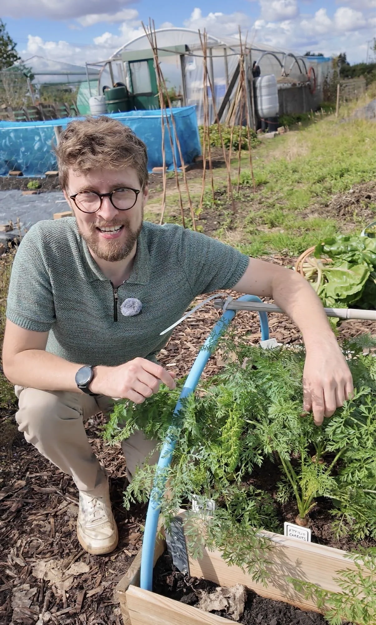 A smiling man wearing glasses and a green short-sleeve shirt, squatting next to a garden bed filled with green plants, holding a cannabis plant, in a backyard garden with a greenhouse and gardening tools in the background.