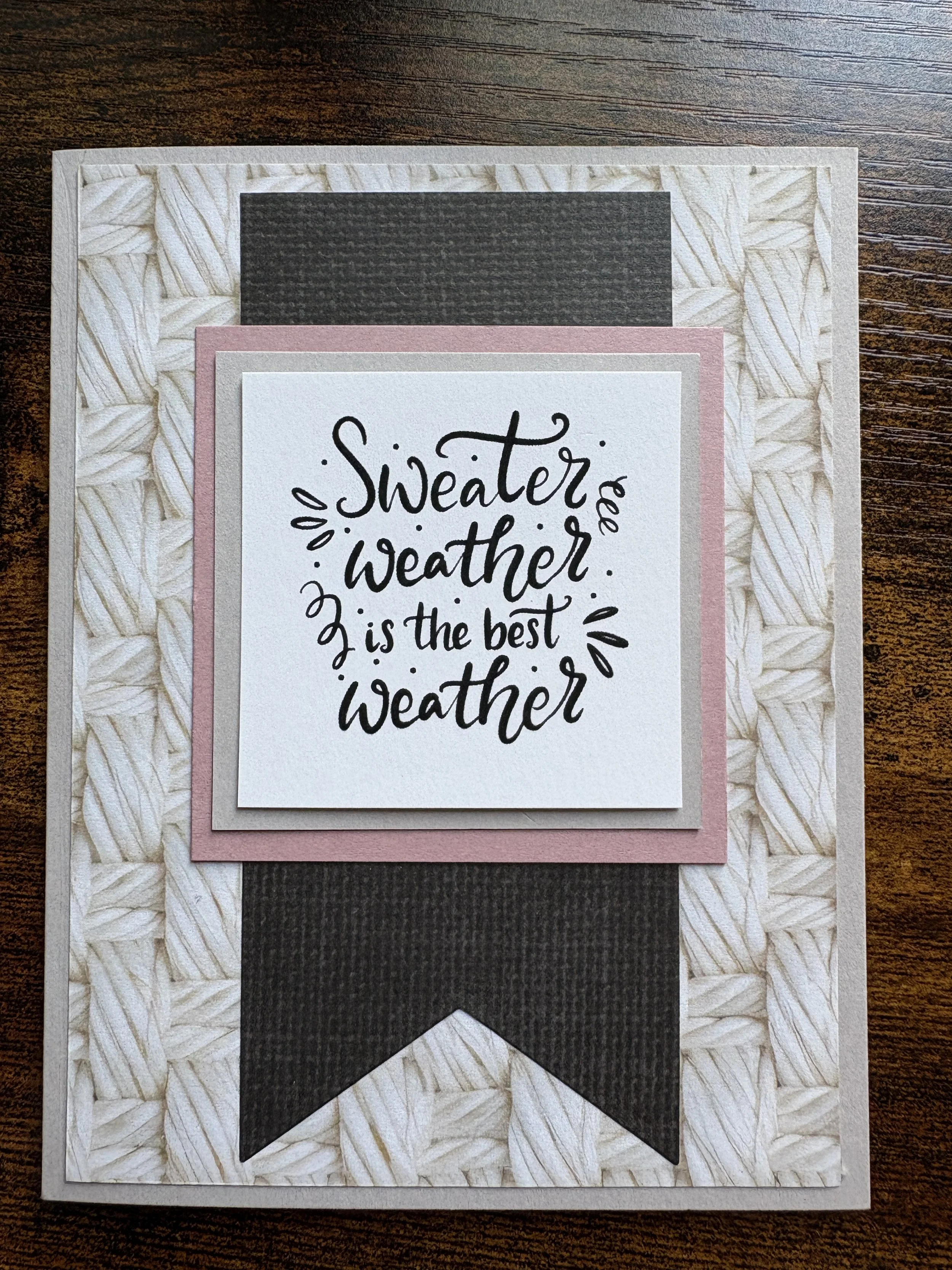 A scrapbook page with layered paper decorations. The top layer features a handwritten quote in black ink: 'Sweater weather is the best weather.' Surrounding it are pink and gray borders, all on a textured beige background, placed on a wooden surface.