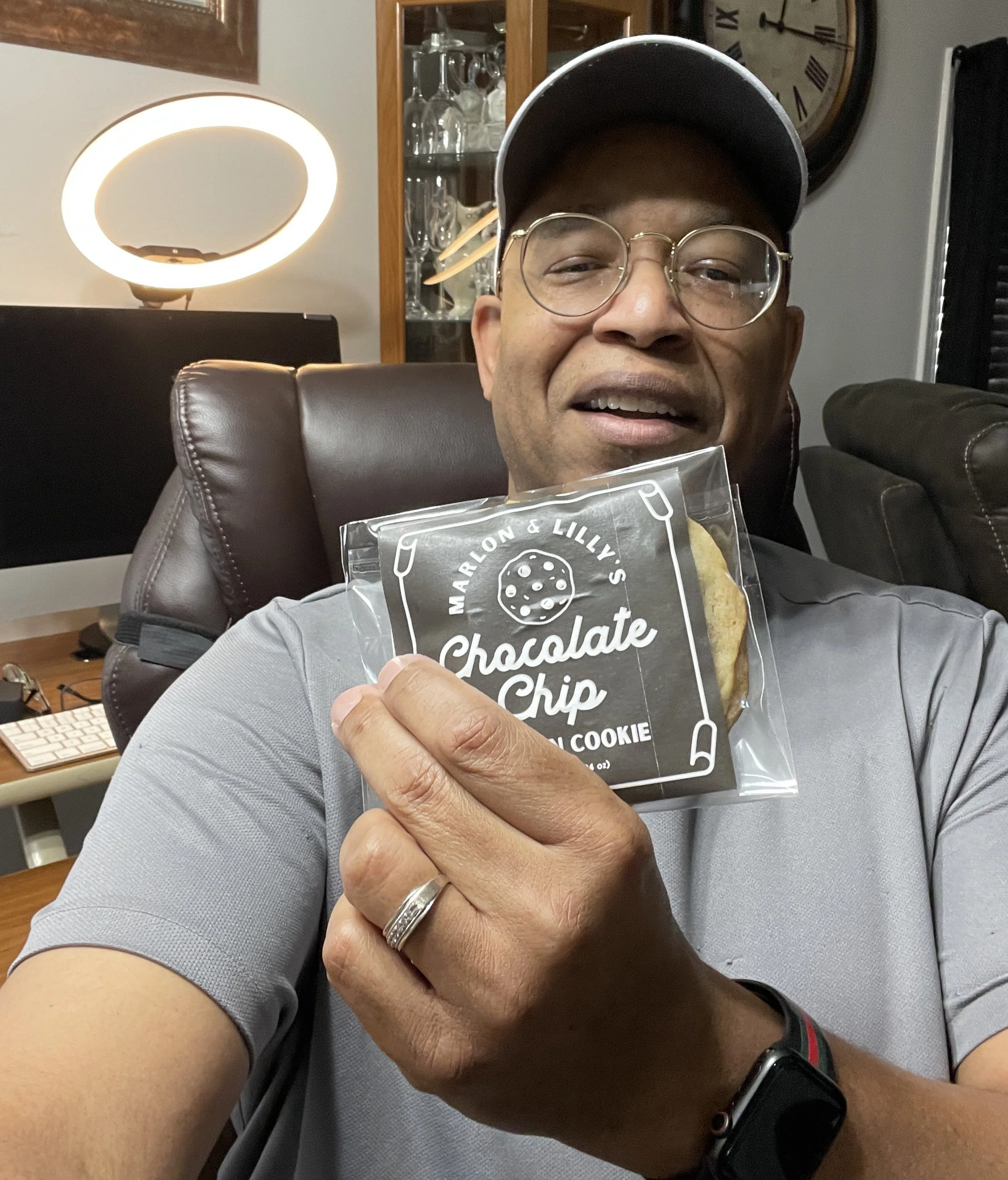 A man wearing glasses and a black cap, holding a package of Marlon & Lily's Chocolate Chip Cookie, sitting in a living room with a clock on the wall, glassware in a cabinet, and a modern lamp in the background.