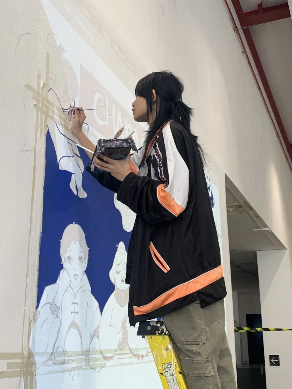 A young woman with black hair painting a mural on a white wall, standing on a yellow ladder, with illustrations of animated characters and text partially visible.