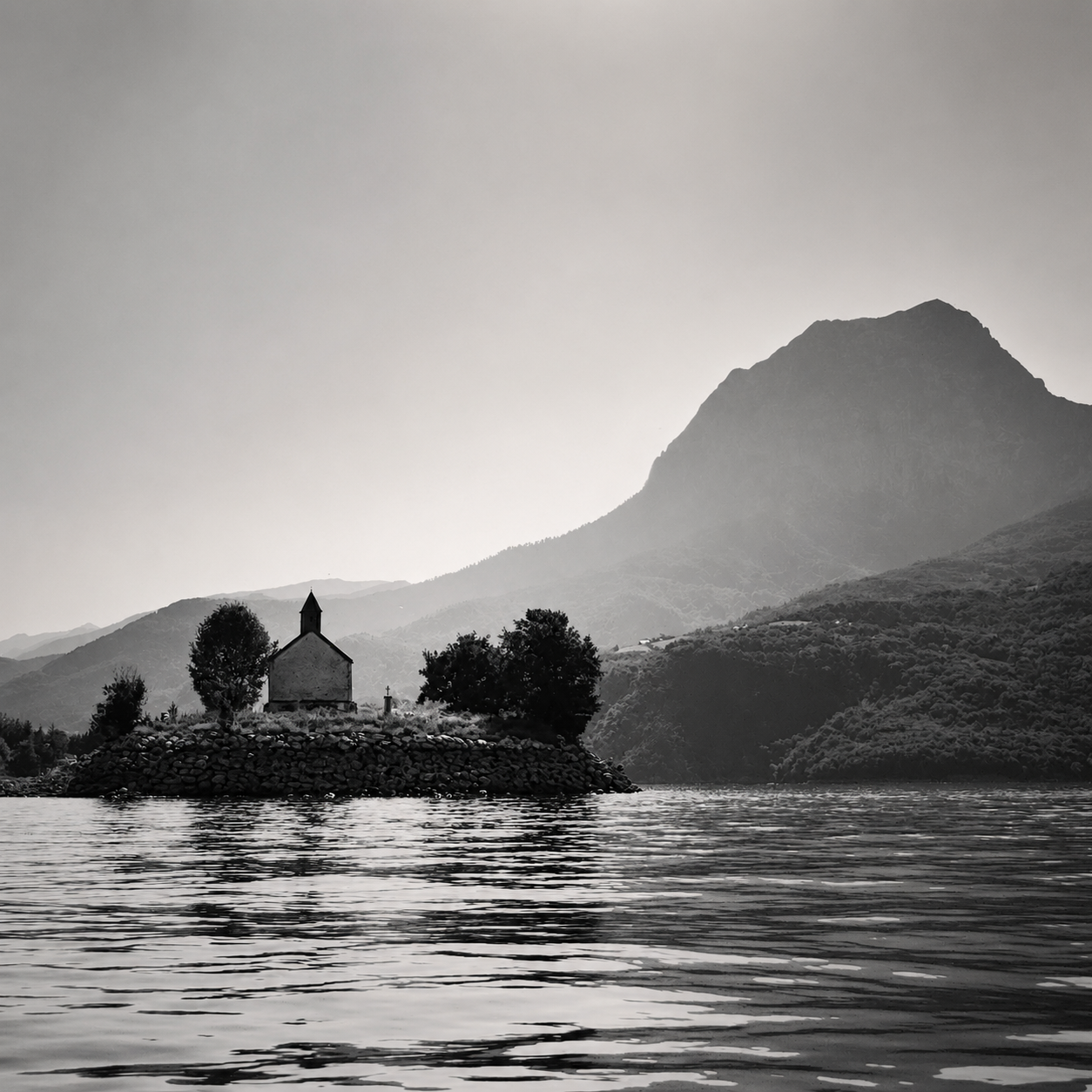 Un paysage en noir et blanc avec un lac, une petite église au centre, entourée d'arbres, avec des montagnes en arrière-plan.
