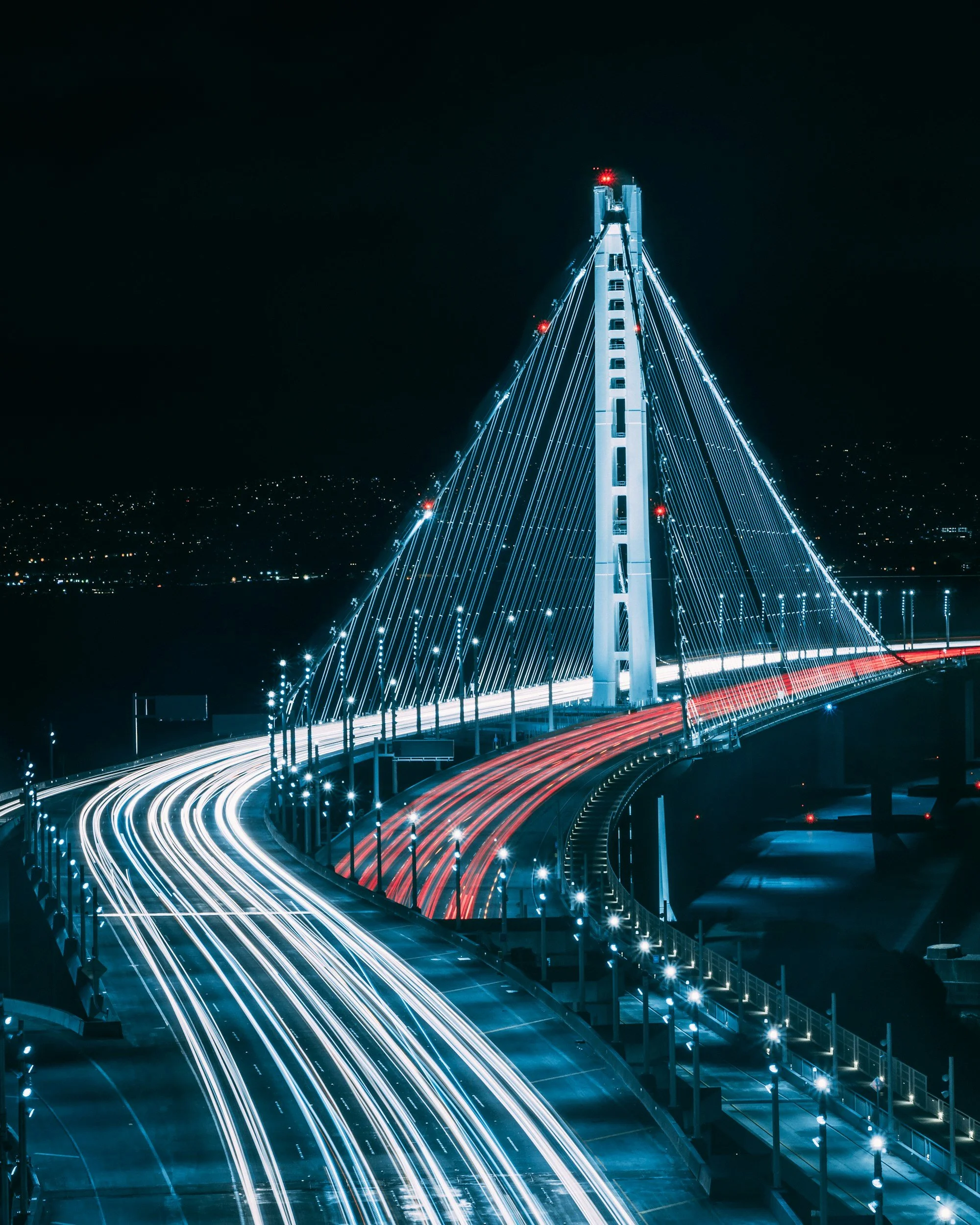 Nighttime view of a modern cable-stayed bridge illuminated with blue lights, with light trails from moving vehicles on the road beneath, and a cityscape in the background.