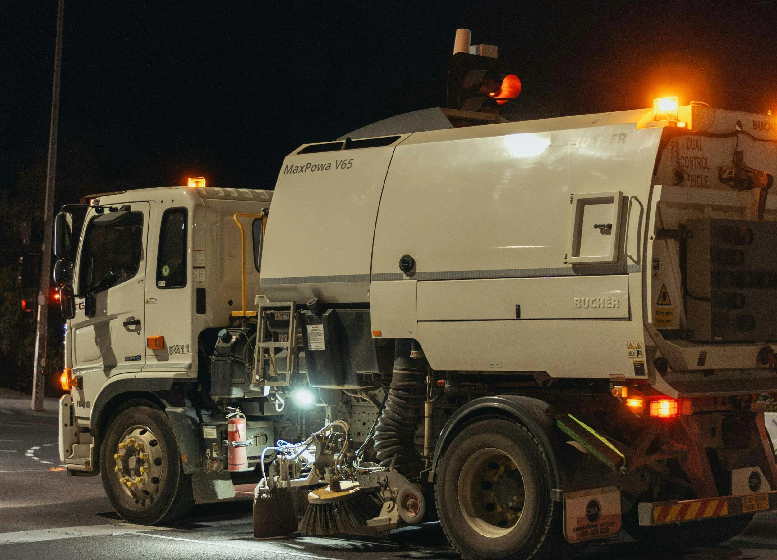 A street sweeper truck parked at night, with orange warning lights flashing, black sky in the background, and a fire extinguisher near the front wheel.