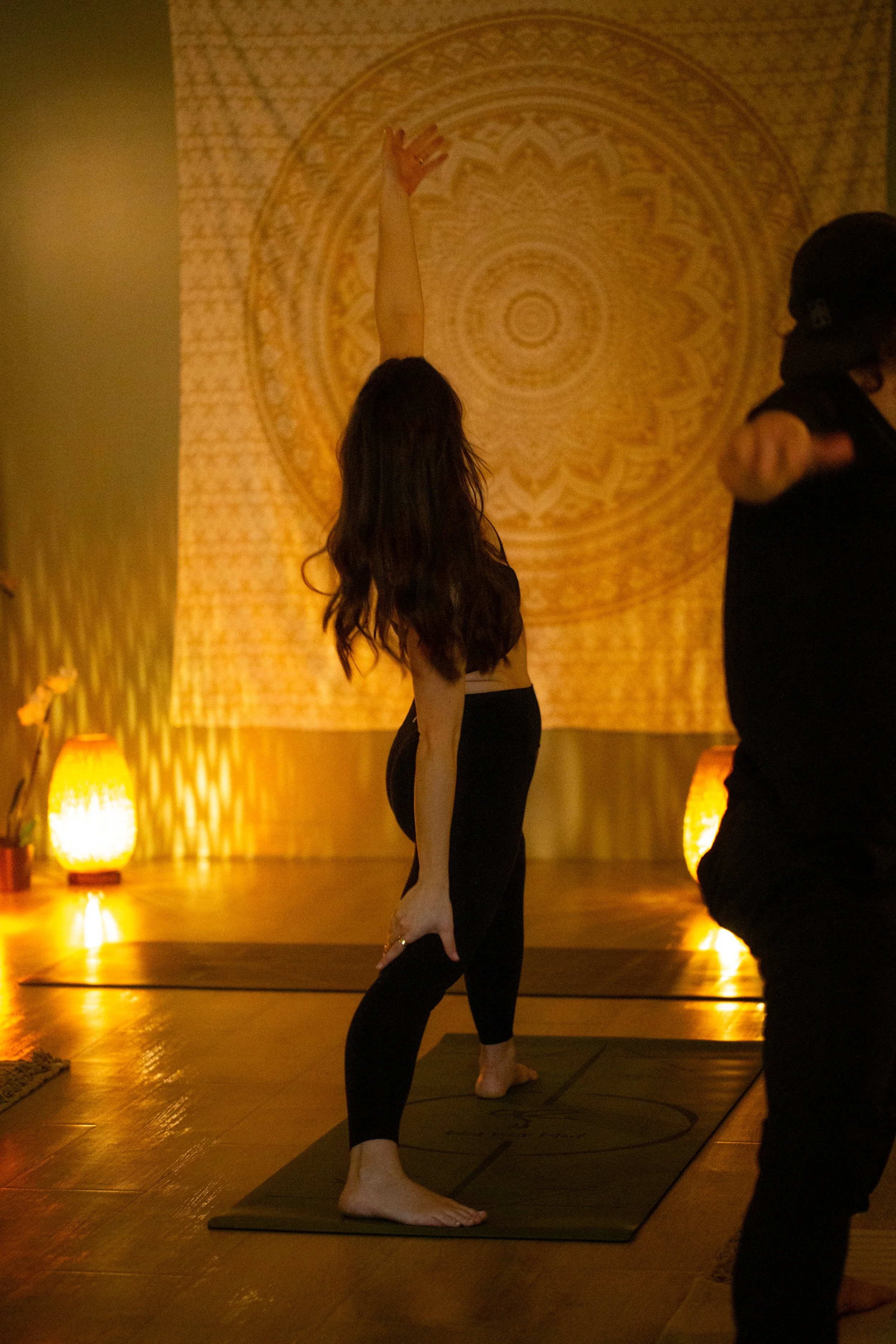 A woman practicing yoga on a mat in a dimly lit room with warm yellow lighting and a decorative mandala on the wall behind her. She is reaching upward with one arm while assistant support her stretched arm.