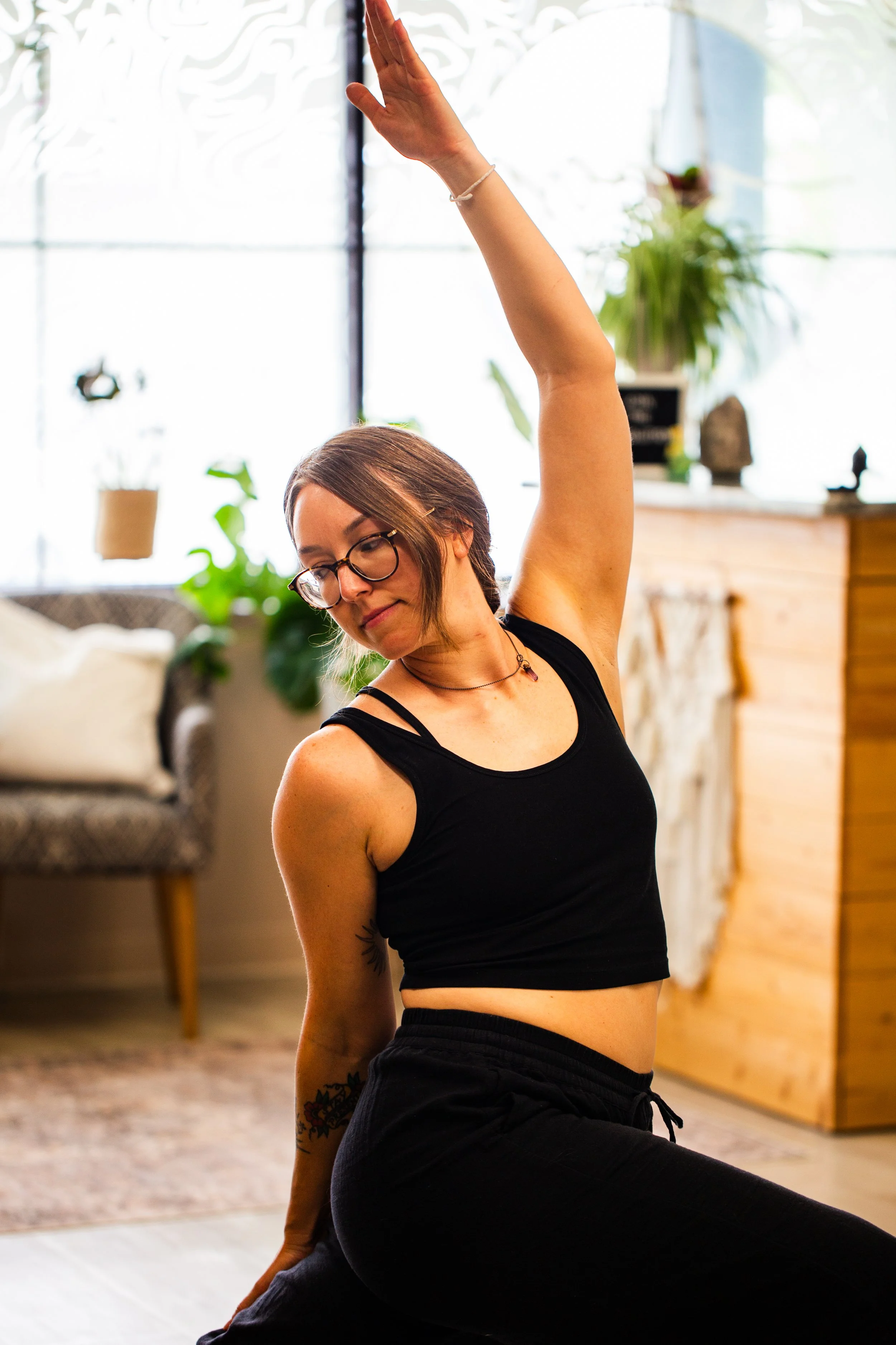 A woman with glasses and tattoos on her arm practicing yoga indoors, wearing a black tank top and black pants, raising her right arm overhead with her eyes closed.