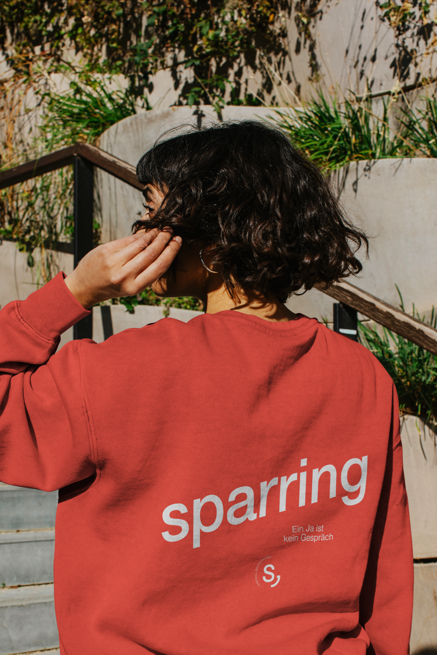 Person mit schwarzen, lockigen Haaren trägt eine rote sweatshirt mit weißen Buchstaben. Hintergrund aus Pflanzen und einer Betonmauer.