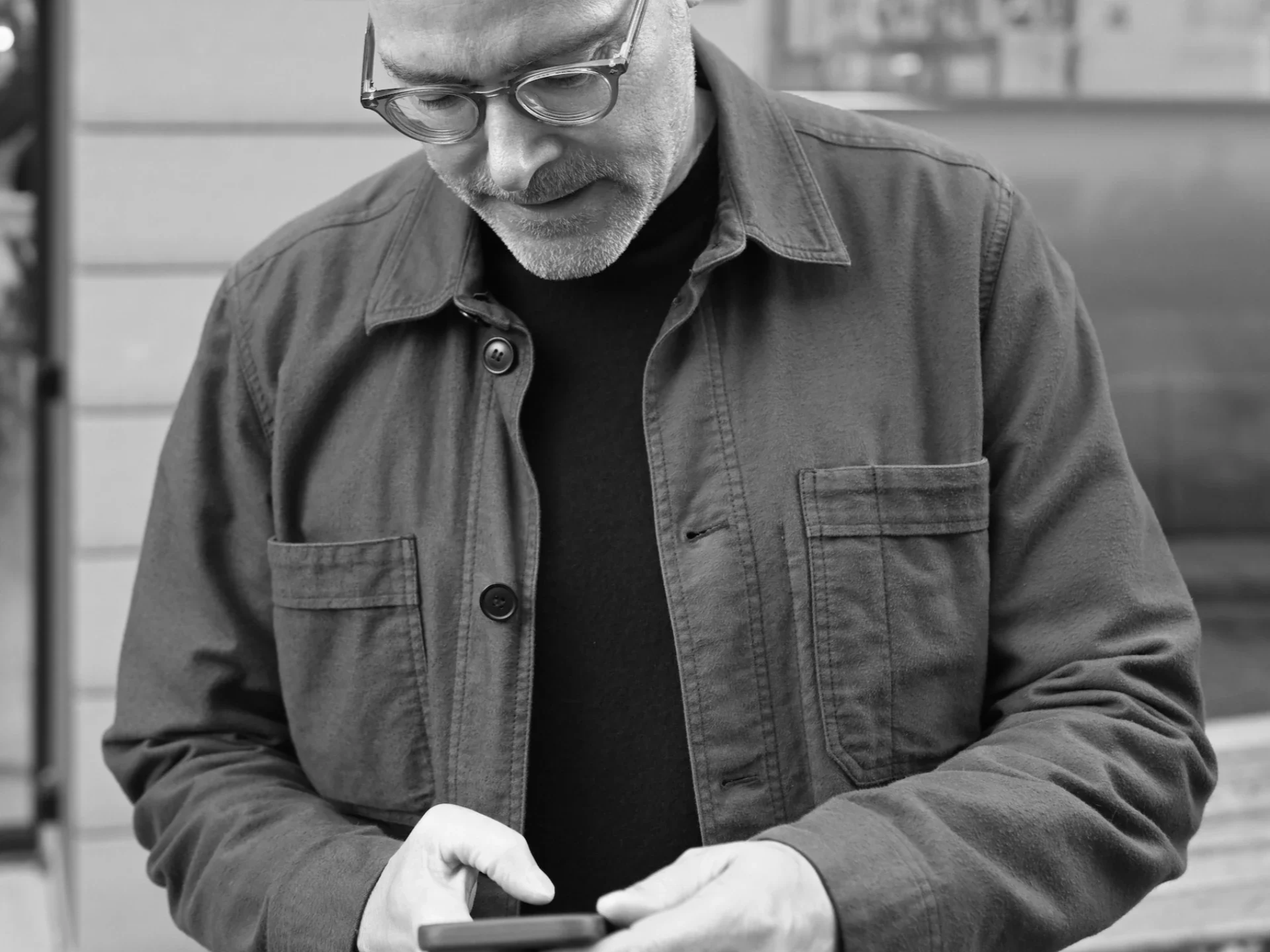 Ein älterer Mann mit Brille trägt eine Jacke und arbeitet mit einem kleinen Tool. Er nutzt ein Smartphone, vermutlich bei der Reparatur oder Prüfung eines Gegenstands.