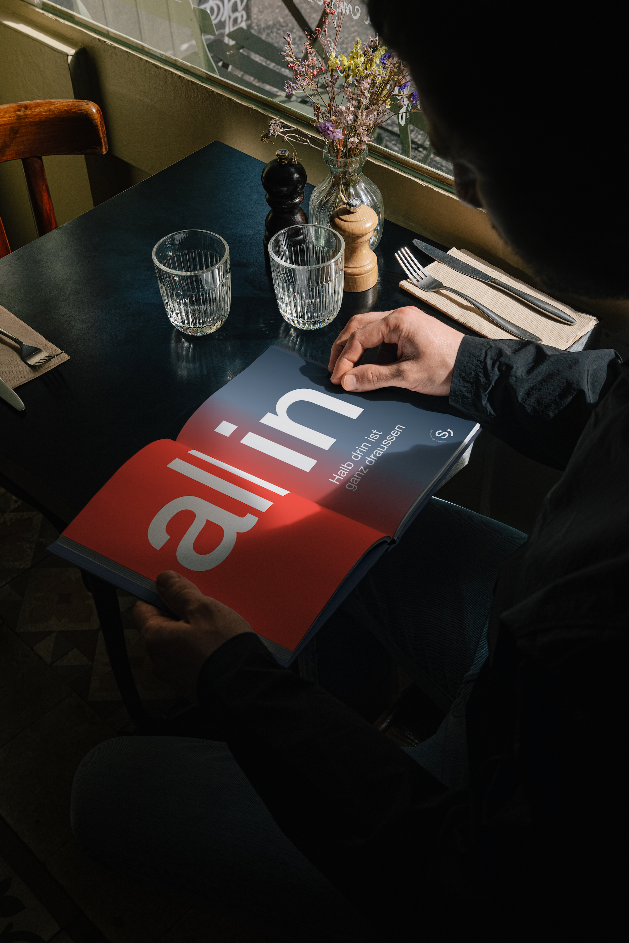 Eine Person liest ein Magazin an einem restauranttisch, auf dem Tisch befinden sich zwei Gläser Wasser, ein Salz- und Pfefferstreuer, eine Pfeffermühle, Messer und Gabeln. Im Hintergrund ist eine Vase mit trockenen Blumen zu sehen.