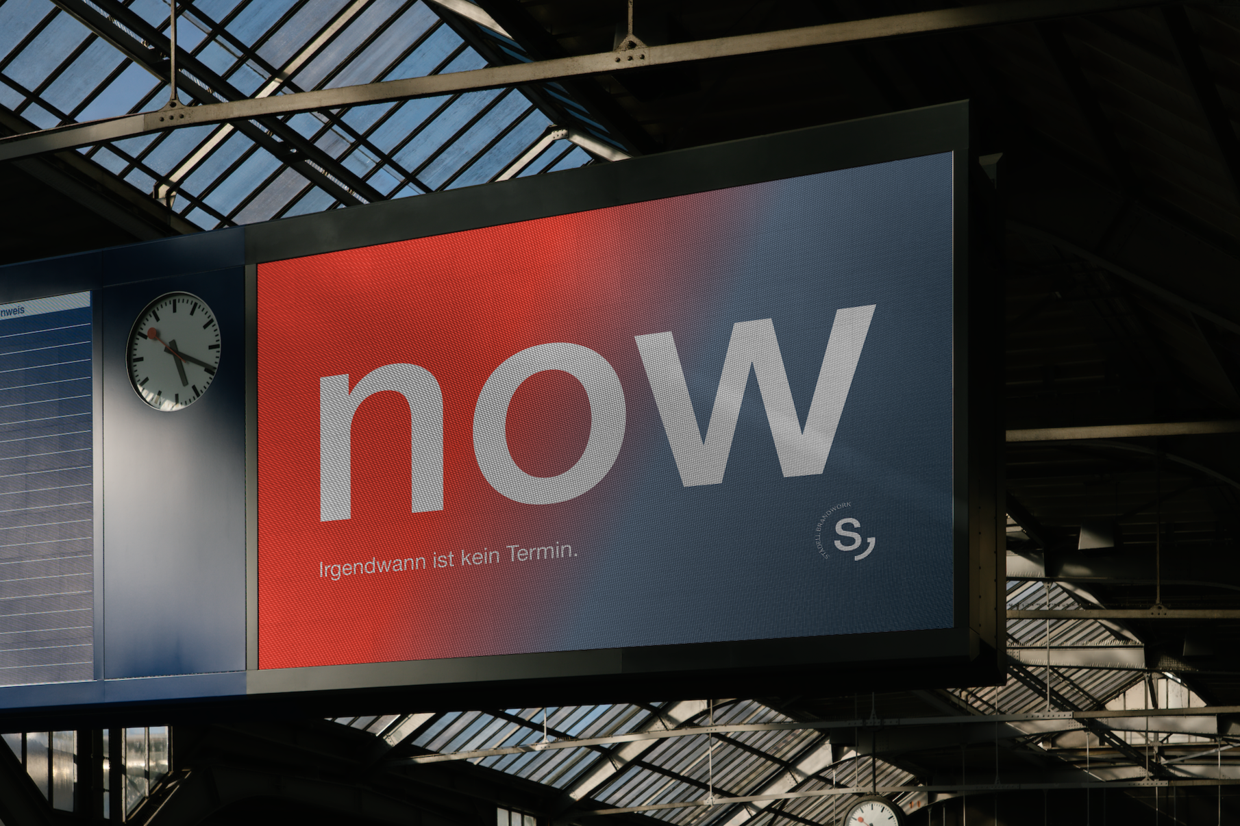 Ein Anzeigetafel mit einem roten Hintergrund mit weißen Buchstaben, die 'now' sagen, und darunter ein kleinerer Text, der lautet 'Irgendwann ist kein Termin'. Im Hintergrund ist ein Glaskupeldach eines Bahnhofs zu sehen sowie eine analoge Uhr.