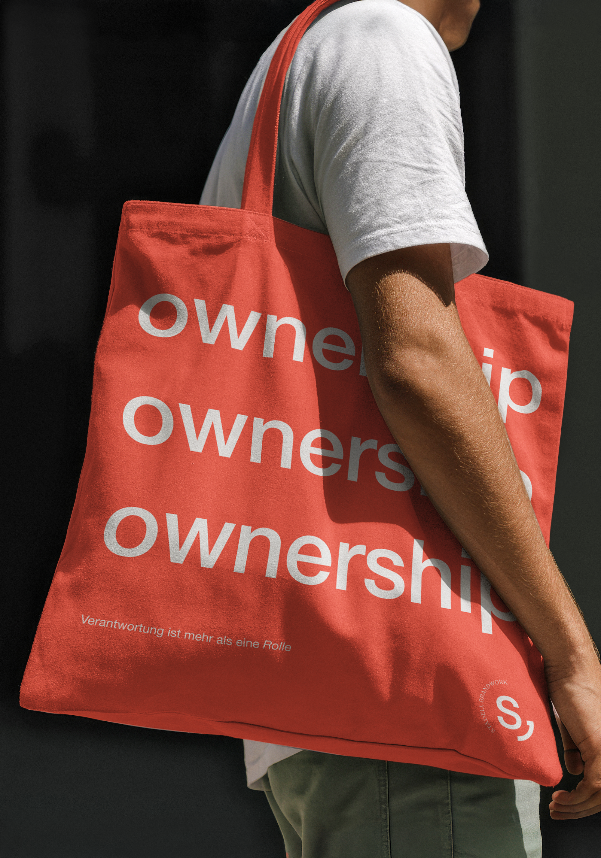 Nahaufnahme eines Menschen mit weißem T-Shirt, der einen roten Stoffbeutel trägt, auf dem das Wort 'Owership' in weißen Buchstaben steht. Der Hintergrund ist dunkel.