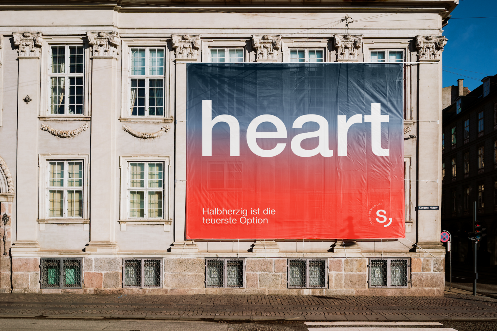 Großes überhängendes Plakat an einer historischen Gebäude-Fassade mit mehreren Fenstern, das den Text 'heart' in weißer Schrift auf einem Farbverlauf von Blau zu Rot zeigt. Darunter steht in kleinerer Schrift 'Halberzlig ist die teuerste Option'.