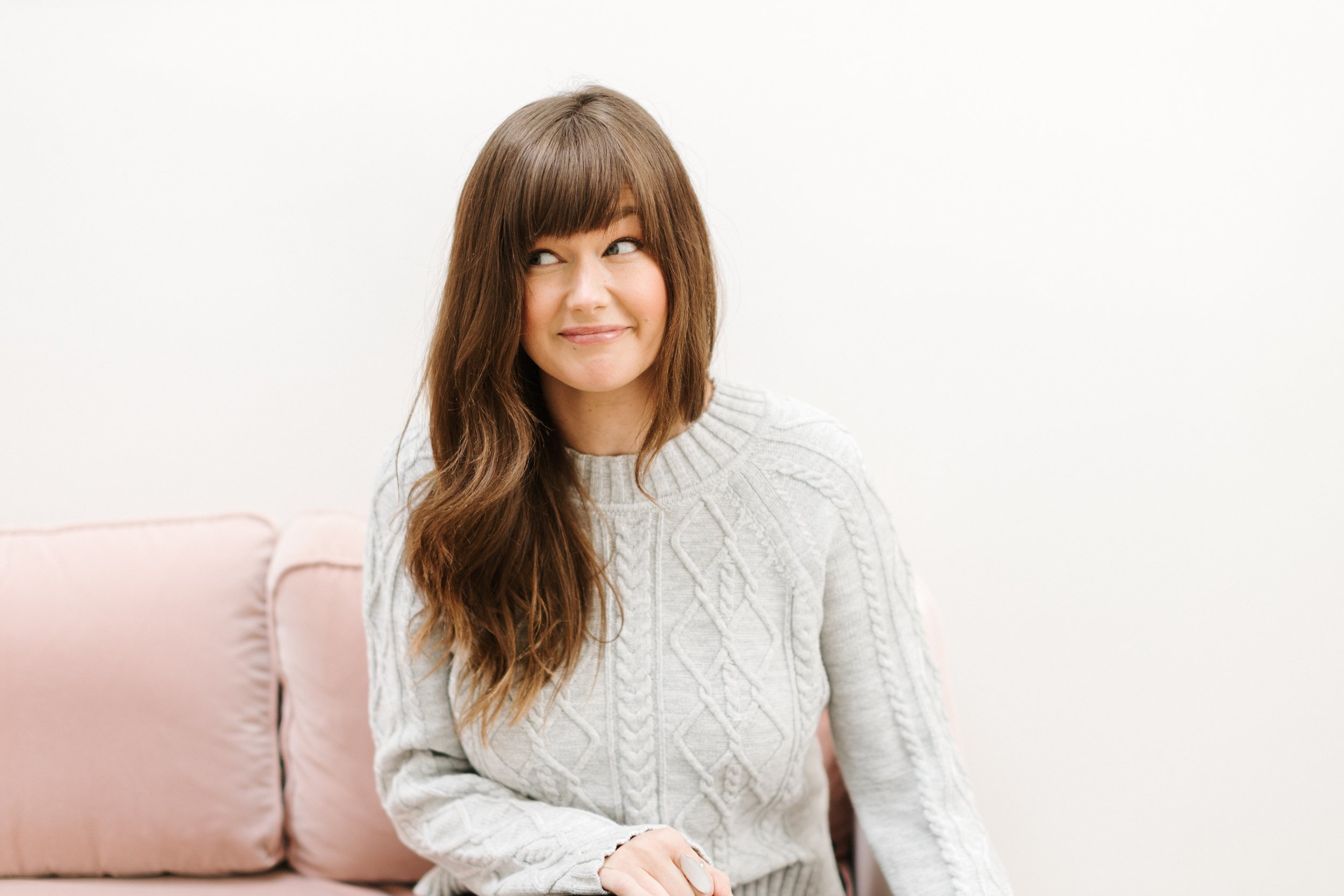 A woman with long brown hair and bangs sitting on a light pink sofa, wearing a gray cable knit sweater, looking to the side with a slight smile, against a white background.