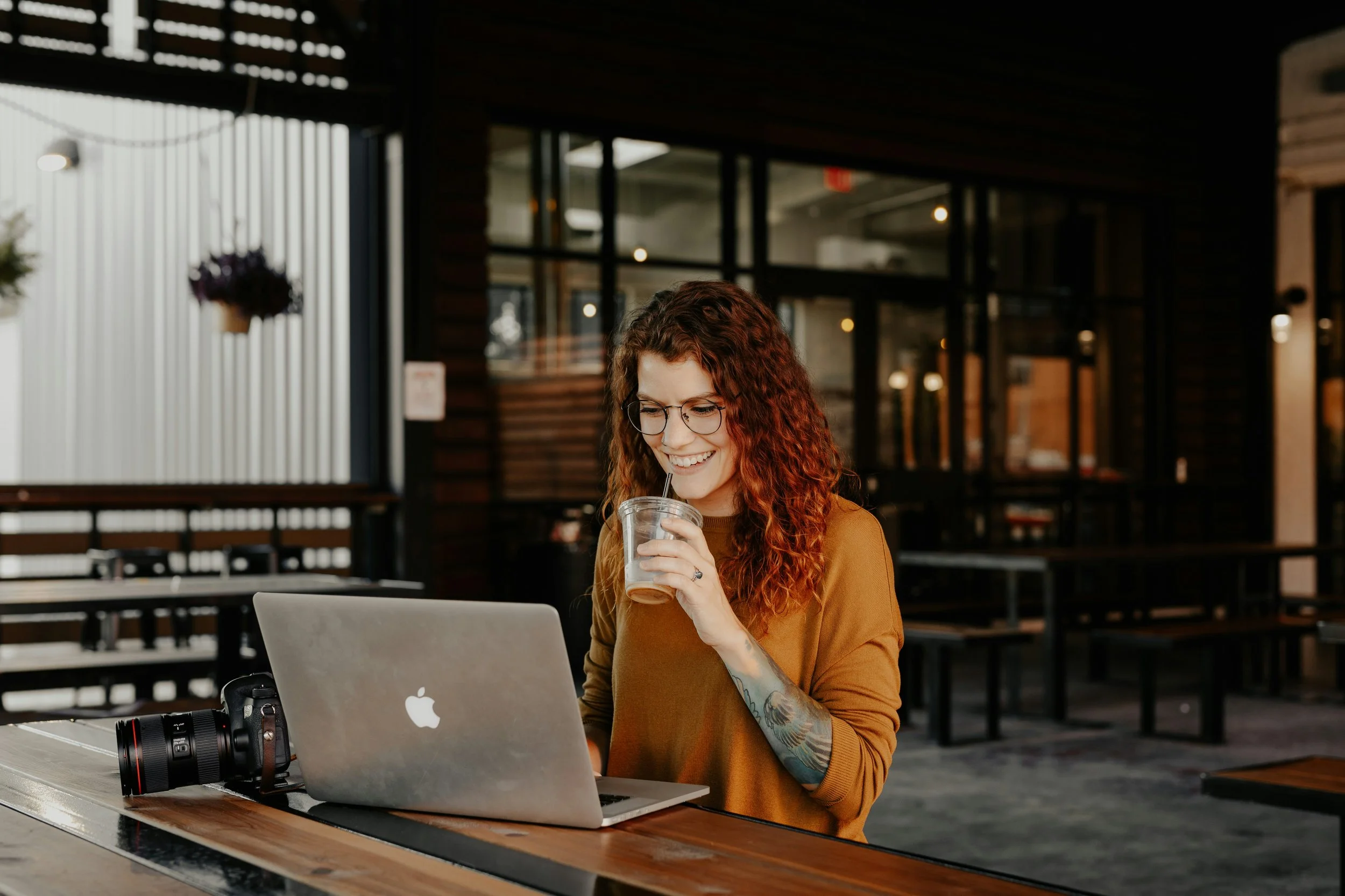 A young woman with curly red hair, wearing glasses and a brown shirt, sitting at a laptop in a modern cafe, drinking from a clear cup with a straw.