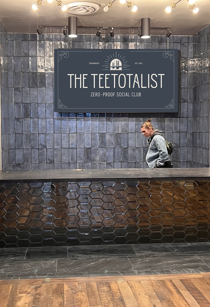 Interior of a social club with a large sign reading 'The Teetotalist Zero-Proof Social Club'. The sign features a logo of a building with rays emanating from it, and the text 'Sacramento' and 'Est. 2022'. There is a woman walking past the reception area, which has a dark, honeycomb-patterned front desk and gray tiled walls.