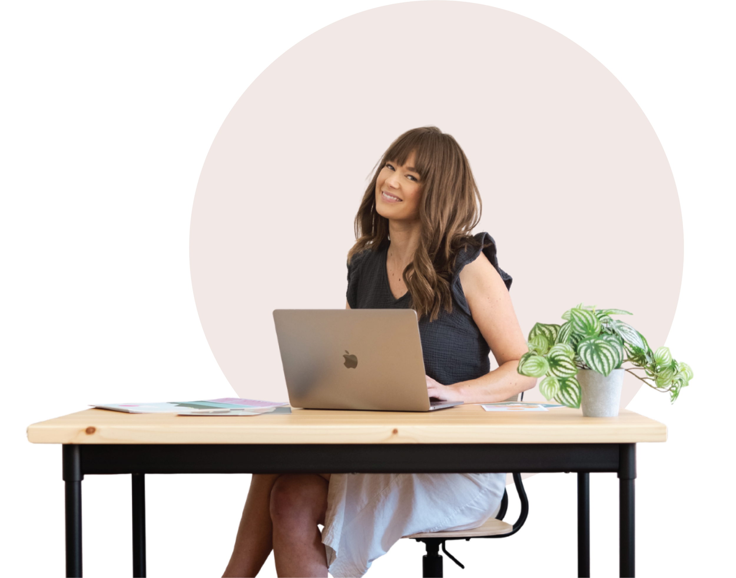 A woman with brown hair and bangs smiling while sitting at a wooden table with a laptop in front of her. There is a potted plant with green leaves on the table, and magazines or papers are spread out in front of her. She is wearing a dark top and a white skirt.